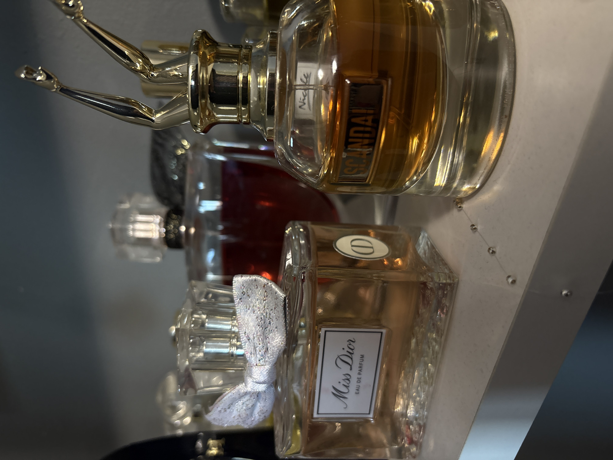 The image shows three perfume bottles on a shelf. The top bottle is "Scandal," with a gold cap shaped like legs. Behind it is a bottle with a red liquid inside. The bottom bottle is "Miss Dior," with a white bow. The photo is taken at an angle, and a fragment of the shelf is visible. The location is not specified in the image, and it's impossible to determine from visual elements alone if it is in Cheshnegirovo, Bulgaria.