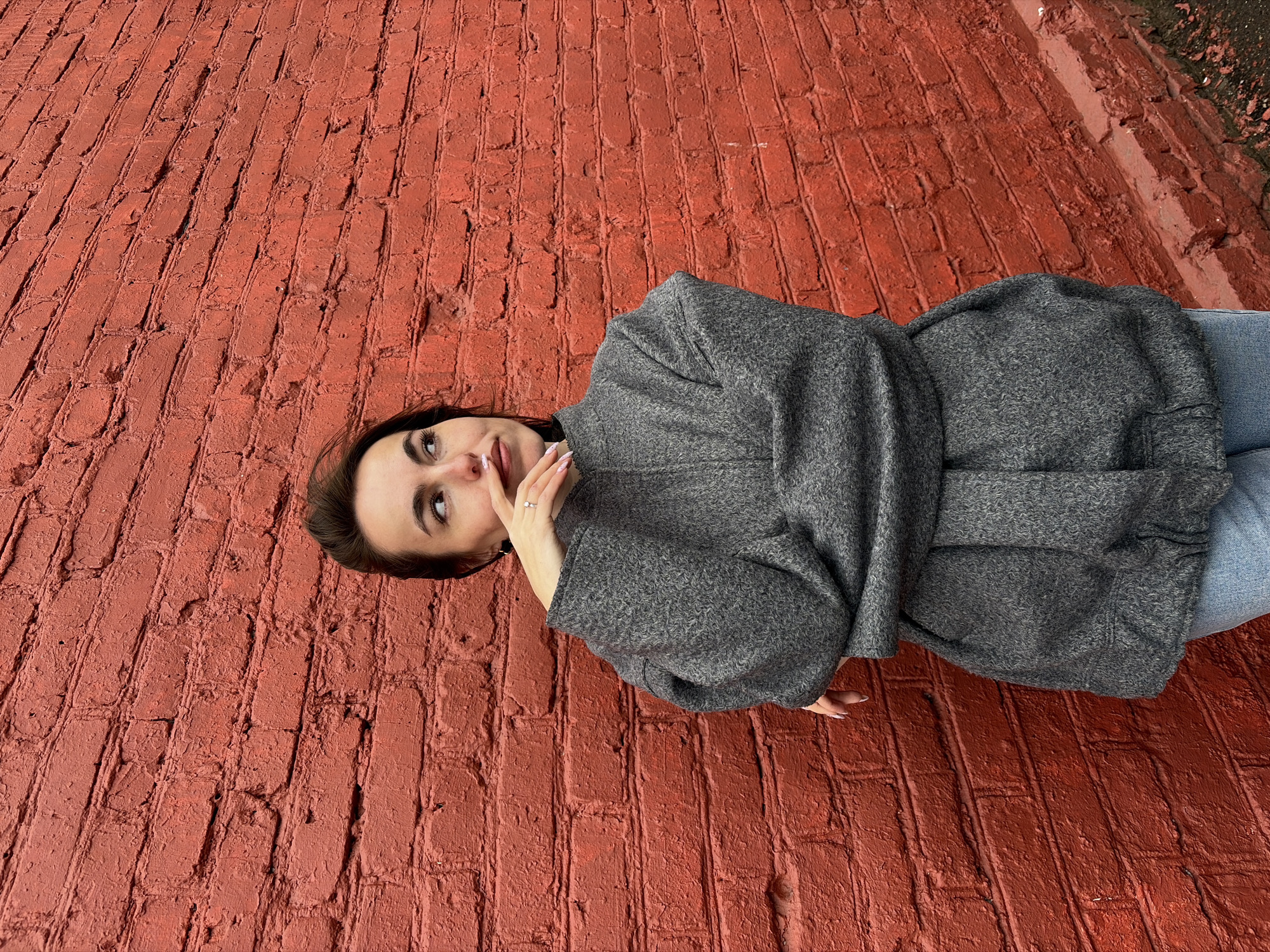 A woman lies on a brick surface. The surface is red and composed of bricks. The woman is wearing a gray coat and blue jeans. Her left hand is near her mouth. She is looking up. The environment is out of focus. It is in Izhevsk, Russia.