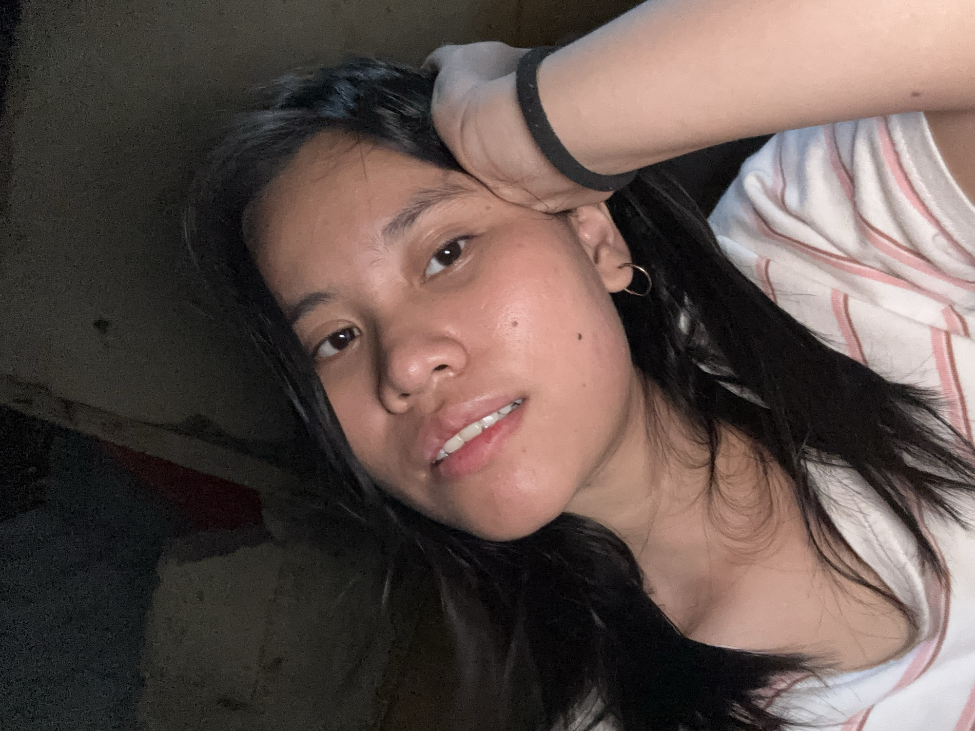 The image is a selfie of a young woman in a dimly lit room. She is resting her head on her right hand, which is adorned with a black wristband. She has dark hair that frames her face and is wearing a striped t-shirt. Her left ear has a small hoop earring. The background is dark and appears to be a wall or furniture. There is no identifiable information about the location in East Jakarta, Indonesia.