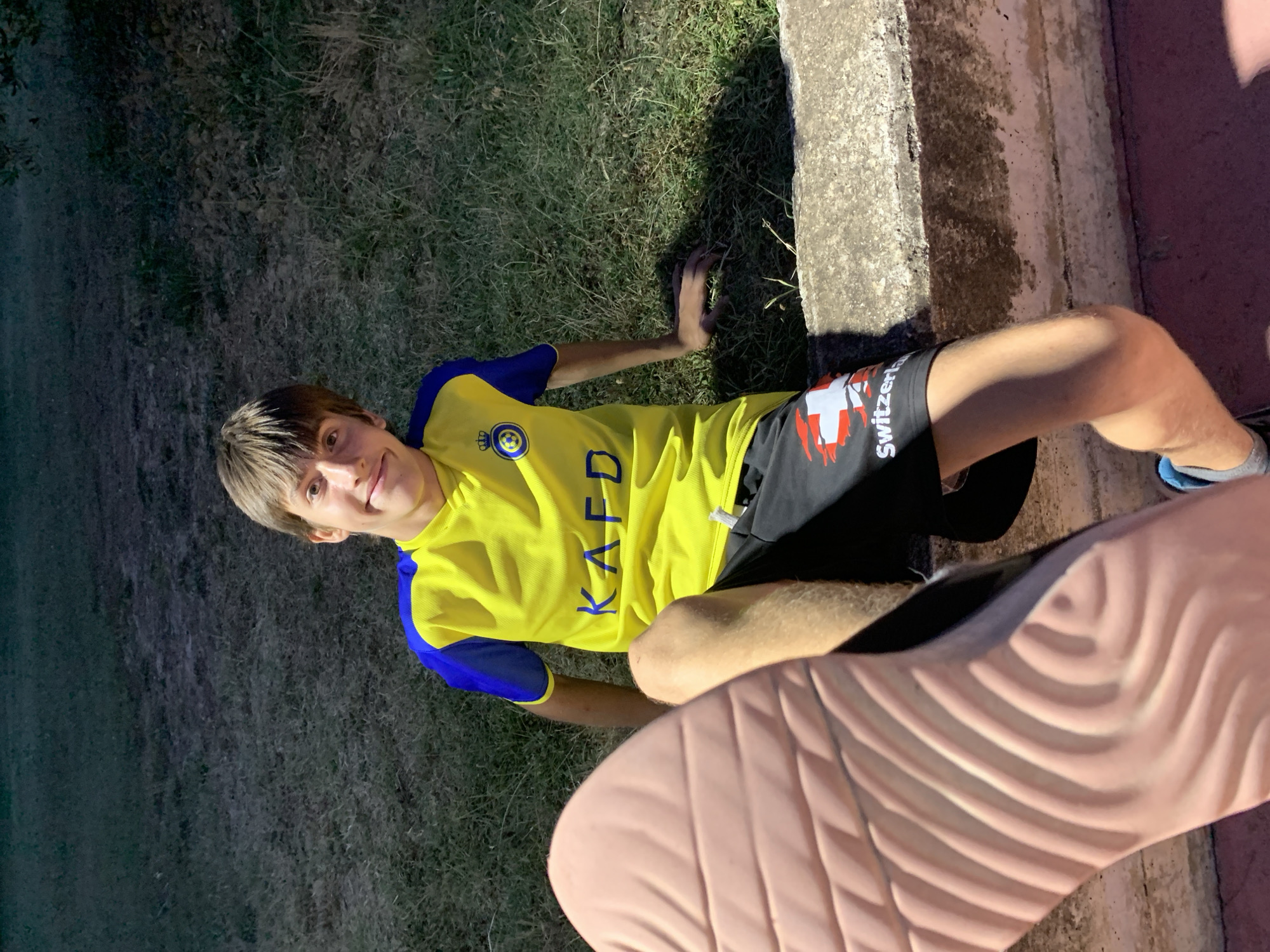 A young person with light brown hair and a bright smile is lying on grass, looking towards the camera. They are wearing a yellow t-shirt with blue sleeves and black shorts with a white Swiss flag emblem and the word "Switzerland" printed on them. The t-shirt has blue lettering that spells "KAFD" and a circular emblem on the left chest. The person is positioned next to a low concrete wall. The scene appears to be outdoors, with dry grass and some shadows suggesting it might be evening or a dimly lit area. A large, textured object, possibly the sole of a shoe or foot, is in the foreground, partially obscuring the lower part of the frame. The overall impression is casual and relaxed. The location is San Quirico d'Orcia, Italy.