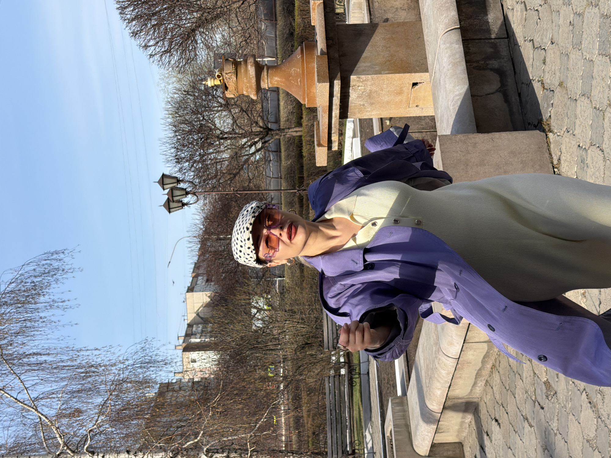 A photograph shows a person standing on the corner of a stone structure near a park in Krasnoyarsk, Russia. The person is wearing a polka dot hat, pink tinted sunglasses, a button-down shirt, and a purple jacket. The park contains trees, benches, and light poles. In the background are apartment buildings. The sky is blue with light clouds.