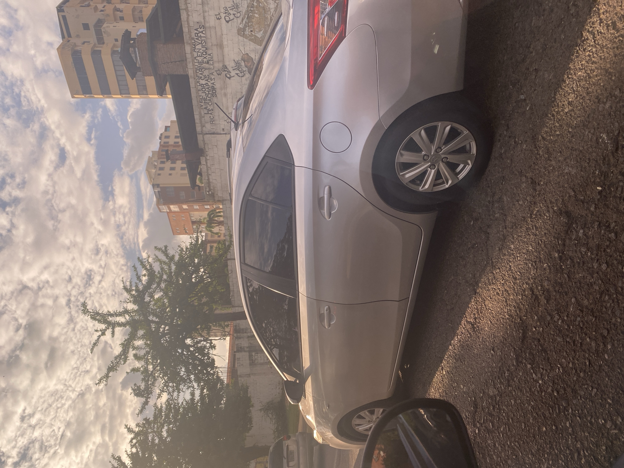 The image depicts a sun-drenched street scene in Maracay, Venezuela, under a partly cloudy sky. The foreground is dominated by a silver sedan parked on a gravel road. The car's right side is fully visible, showing its doors, windows, and rear wheel with a silver alloy rim. A small portion of another vehicle's side mirror is visible in the lower left corner, suggesting the viewer is inside another car.
In the background, a low concrete wall displays graffiti, with some text that appears to be advertisements or names. Behind the wall, several trees with dense foliage are present, and beyond them, urban buildings rise into the sky. Two notable buildings are visible: one is a tall, yellowish structure with multiple floors and dark-tinted windows, and another is a shorter, multi-story building with a reddish-orange facade. The overall lighting suggests it's either early morning or late afternoon due to the warm, angled sunlight casting shadows. The environment appears to be an urban or semi-urban area within Maracay.