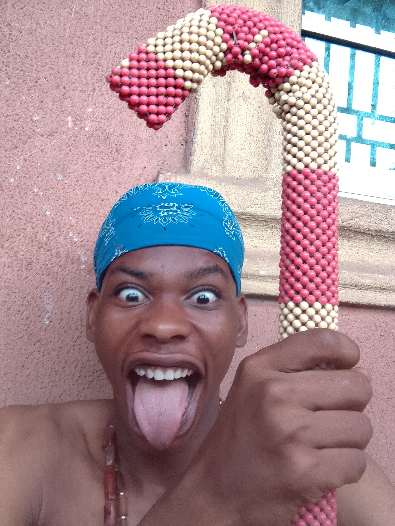 The photograph depicts a man with dark skin, wearing a blue bandana with white patterns, making a face with wide eyes and tongue sticking out. He is holding a beaded cane, patterned with alternating red and cream-colored beads. The background consists of a peach-colored wall and a window with a white grid pattern. The location is the city of Unknown, Unknown. The man is wearing a necklace with red and brown beads. The man appears to be posing playfully for the camera.