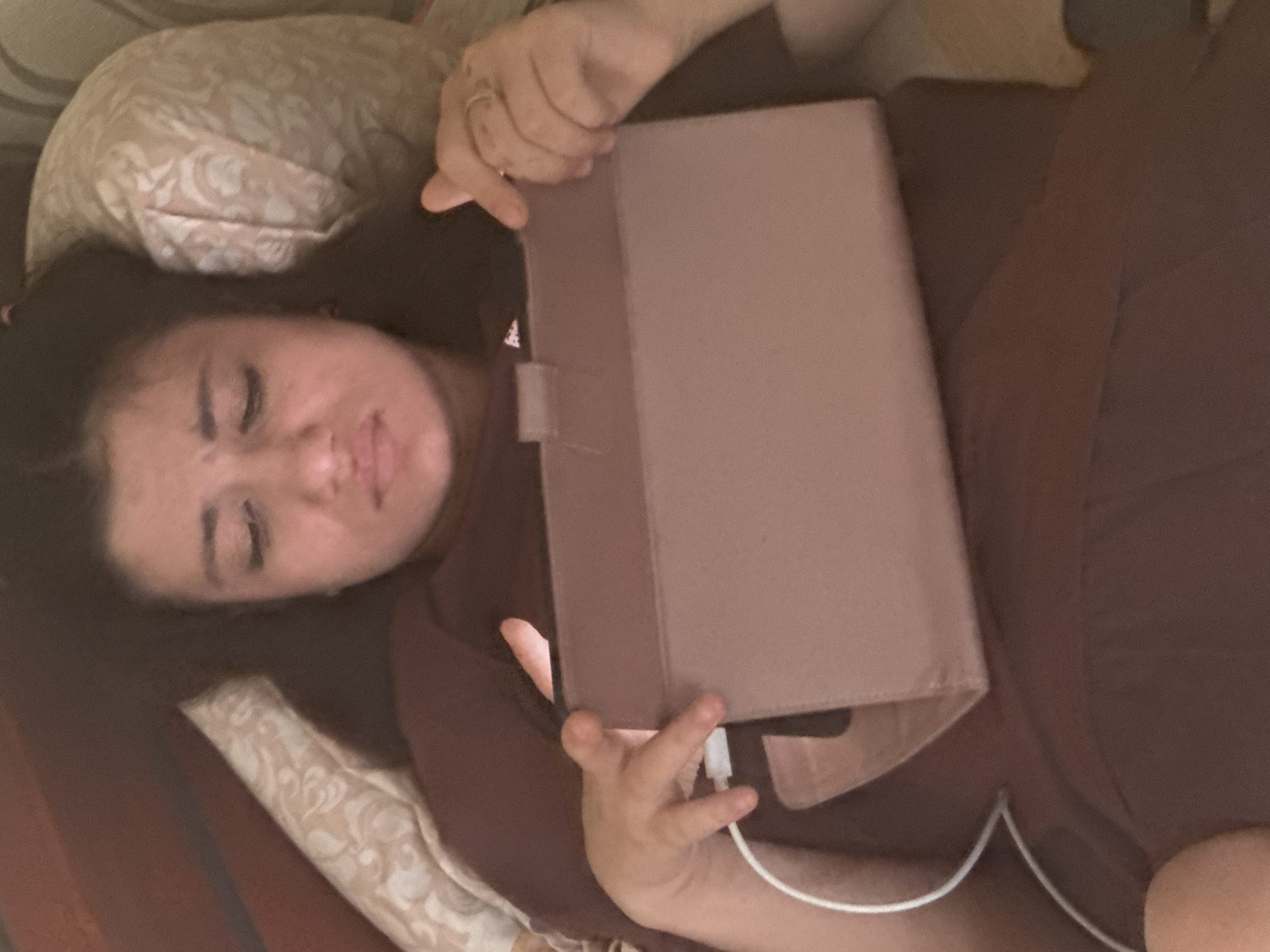 An adult female with dark hair is depicted lying supine on a surface, likely a bed, within an indoor environment. She is wearing a dark brown, short-sleeved top. Her head rests against a light-colored pillow featuring a subtle, patterned fabric. She holds a rectangular electronic tablet, encased in a reddish-brown folio cover, in front of her. Her left hand positions the tablet, with a white charging cable visibly connected to its left side. Her right hand rests on the tablet's right side, adorned with a ring on the ring finger. The individual's eyes are cast downwards, focused on the tablet screen. The overall lighting is subdued, suggesting a low-light condition, with the tablet screen emitting light onto her face and hands. A dark brown, rigid structure, possibly a headboard, is visible along the left edge of the frame. The location is in the city of Unknown, Unknown.