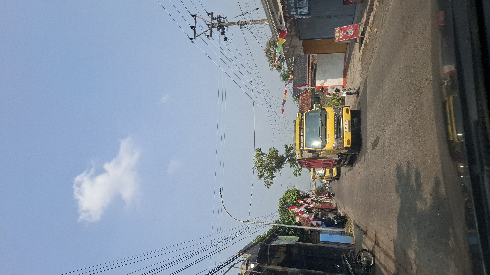 The image depicts a street scene in Sukasari, Indonesia, on a clear day with a blue sky and scattered white clouds. The road is paved and appears to be a two-lane street. Several yellow trucks are present, with the most prominent one in the foreground displaying the license plate "F 8729 VC". The trucks are traveling on the road, with one person standing on the side of the road next to a barber shop sign that reads "PANEKAS RAMBUT BARBER".
On the left side of the road, there are buildings with awnings and several Indonesian flags are displayed, suggesting a local celebration or event. Motorcycles are parked along the sidewalk on the left. Overhead, power lines crisscross the sky, supported by utility poles. On the right side, there are more buildings and flags. The overall impression is of a busy, somewhat narrow street in a residential or commercial area within a town or city.