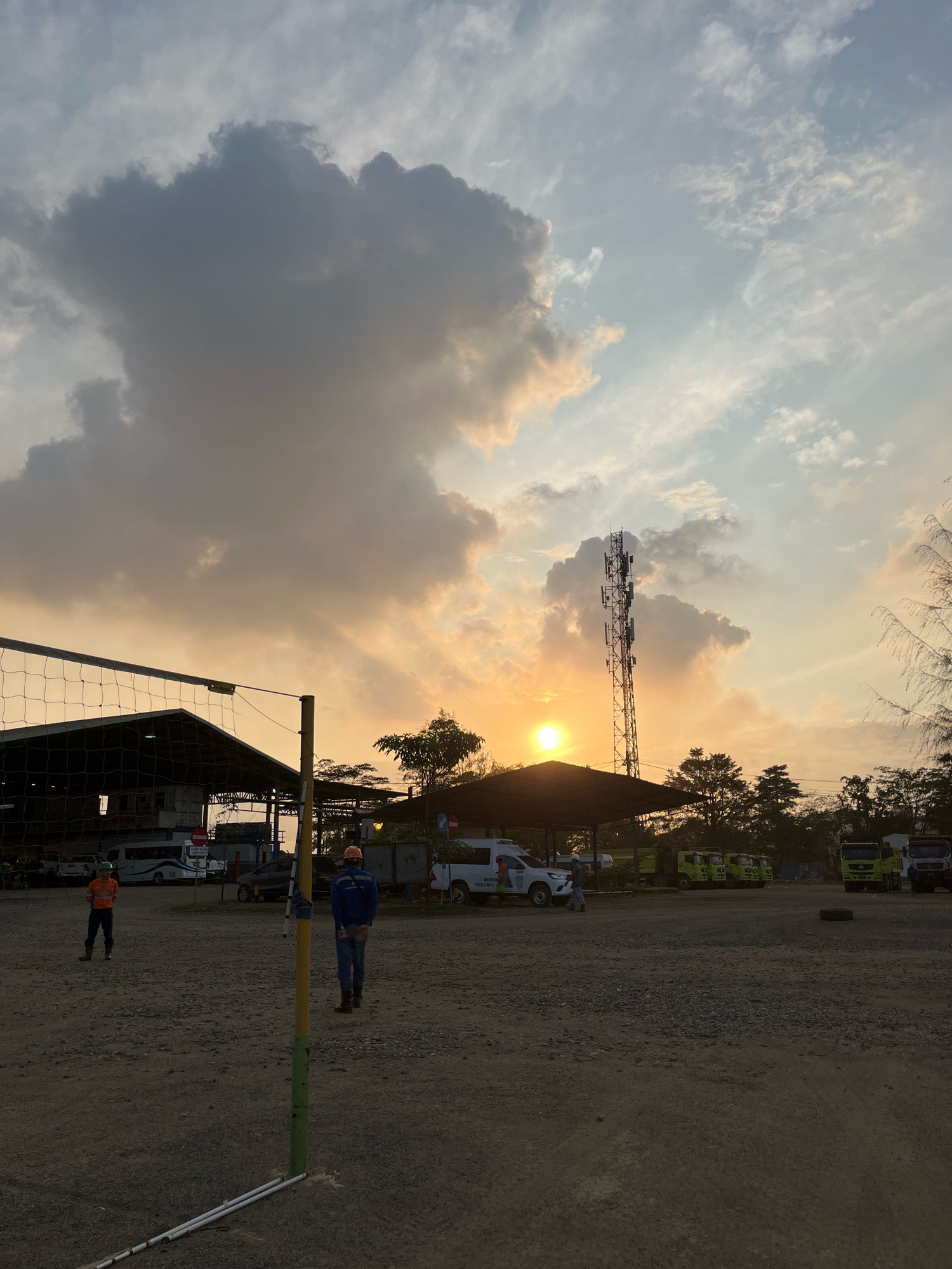 In an outdoor, dusty lot, two individuals are present near a volleyball net. One person, wearing an orange shirt and a hard hat, stands to the left, facing the net. The second person, in a blue jacket and jeans, wearing an orange hard hat, stands to the right, also facing the net. The setting appears to be an industrial area with a large building or warehouse structure in the background, partially covered by a roof. Several vehicles are parked, including a white van with text that reads "MORON SEKUTRIADE", and multiple green trucks. A cell tower is visible in the distance. The sky is illuminated by a sunset, with dramatic clouds casting long shadows across the scene. The overall impression is a functional, possibly work-related, outdoor space at dusk.