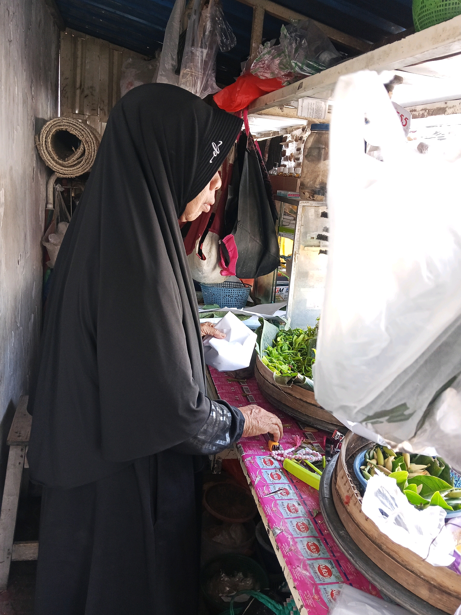 The image depicts an elderly woman in a black hijab and a dark abaya at a market stall in Wates, Indonesia. She is standing behind a table laden with goods. To her left, a rolled-up mat is propped against a wall. On the table, there are packets of what appear to be antiseptic wipes, small pink and green rosary beads, a yellow plastic tool, and a small orange object. In front of her, a woven circular container holds green leafy vegetables and some white items, possibly flowers or buds. A clear plastic bag obscures part of the scene to the right. The stall is sheltered by a blue corrugated metal roof, and various items, including red plastic bags, are hanging from the ceiling. The wall to the left is rough and textured. The woman's hands are visible as she appears to be arranging or selecting items on the table. The overall impression is that of a humble, traditional market setting.