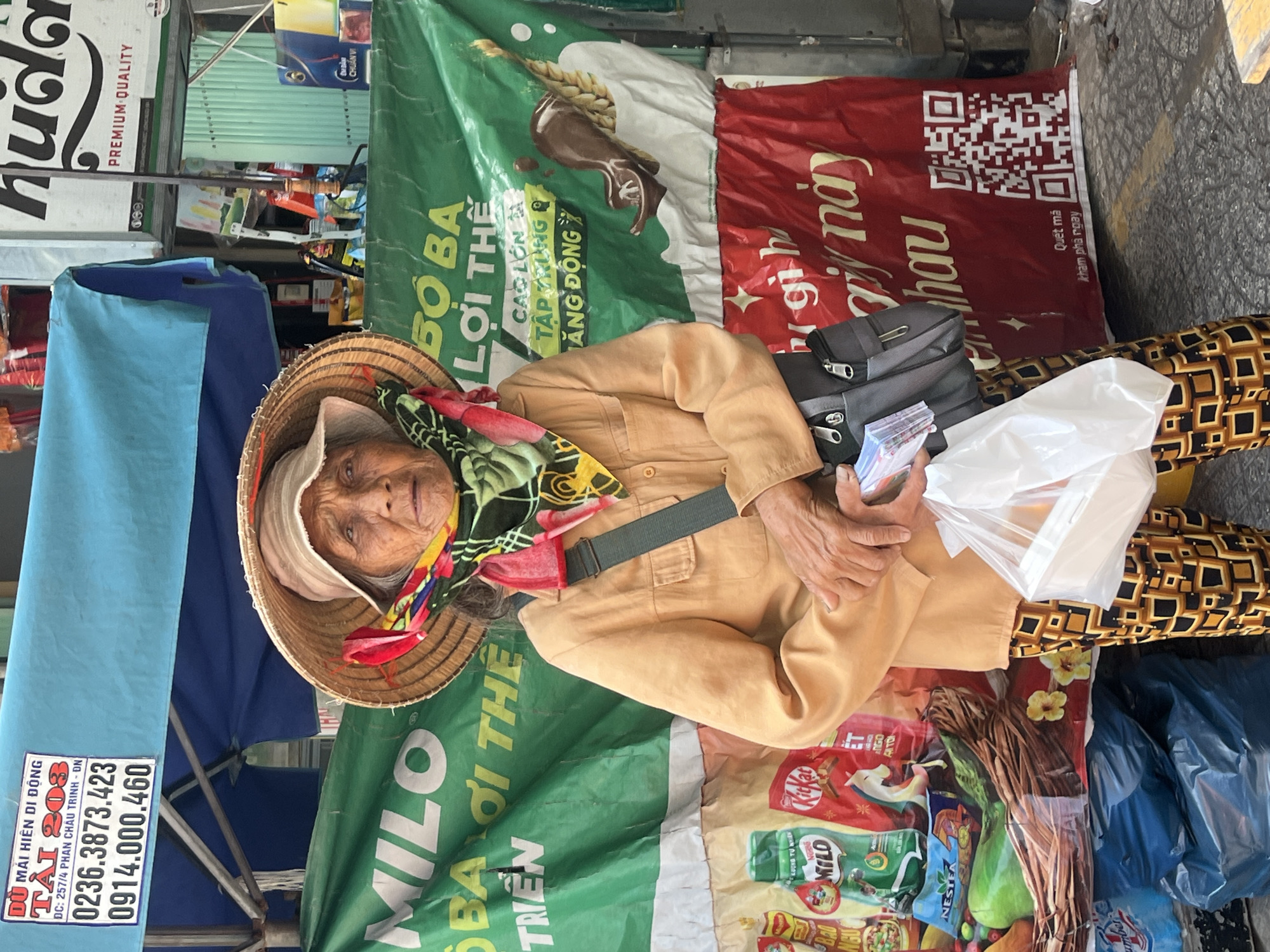 An elderly Vietnamese woman, wearing a conical hat and a colorful scarf, stands in front of a stall in Da Nang, Vietnam. She is dressed in a tan shirt and patterned pants. In her hands, she holds a white plastic bag and some papers. A dark gray shoulder bag is slung across her body. The stall is decorated with a large green banner featuring images of various food products, including Milo and Kit Kat, and a red banner with Vietnamese text and a QR code. The background shows a blue canopy and a light-colored building. The overall scene suggests a street market or food stall in a busy urban environment.