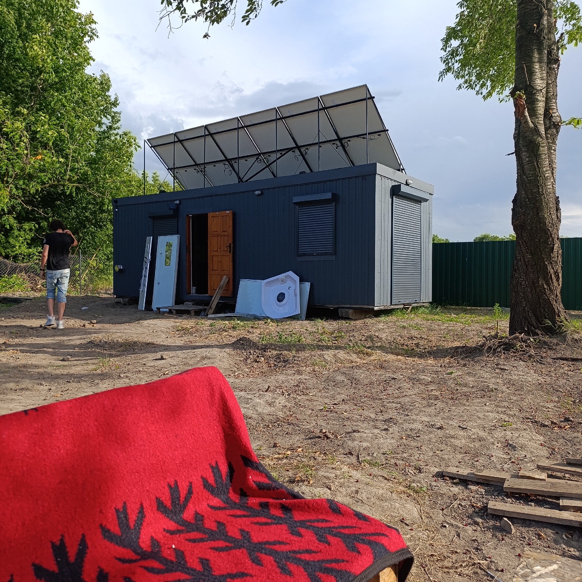 In Ladyzhyn, Ukraine, a dark grey shipping container, repurposed as a dwelling, is situated outdoors on a patch of uneven, sandy ground. The container is equipped with solar panels mounted on its roof, angled upwards. Adjacent to the container, a person in a black t-shirt and light blue denim shorts stands with their back to the camera, facing away from the dwelling. Outside the container, various objects are present: two doors leaning against the side, a white bathtub or basin, and wooden planks scattered on the ground. The scene is partially framed by trees and a green metal fence. In the foreground, a red blanket with a black leaf-like pattern is draped over an unseen object. The sky is partly cloudy.