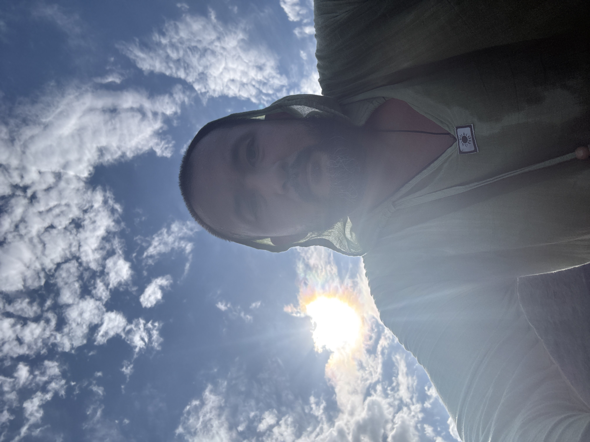 The image is a selfie taken outdoors under a bright, partly cloudy sky. The sun is shining intensely, creating a lens flare. The subject, a man with a beard and wearing a light green hooded garment, is looking at the camera. He is wearing a necklace with a pendant that appears to have a sun symbol on it. The background consists of a blue sky with scattered white clouds. There are no identifiable landmarks that suggest the location in the city of Ақши, Kazakhstan.
