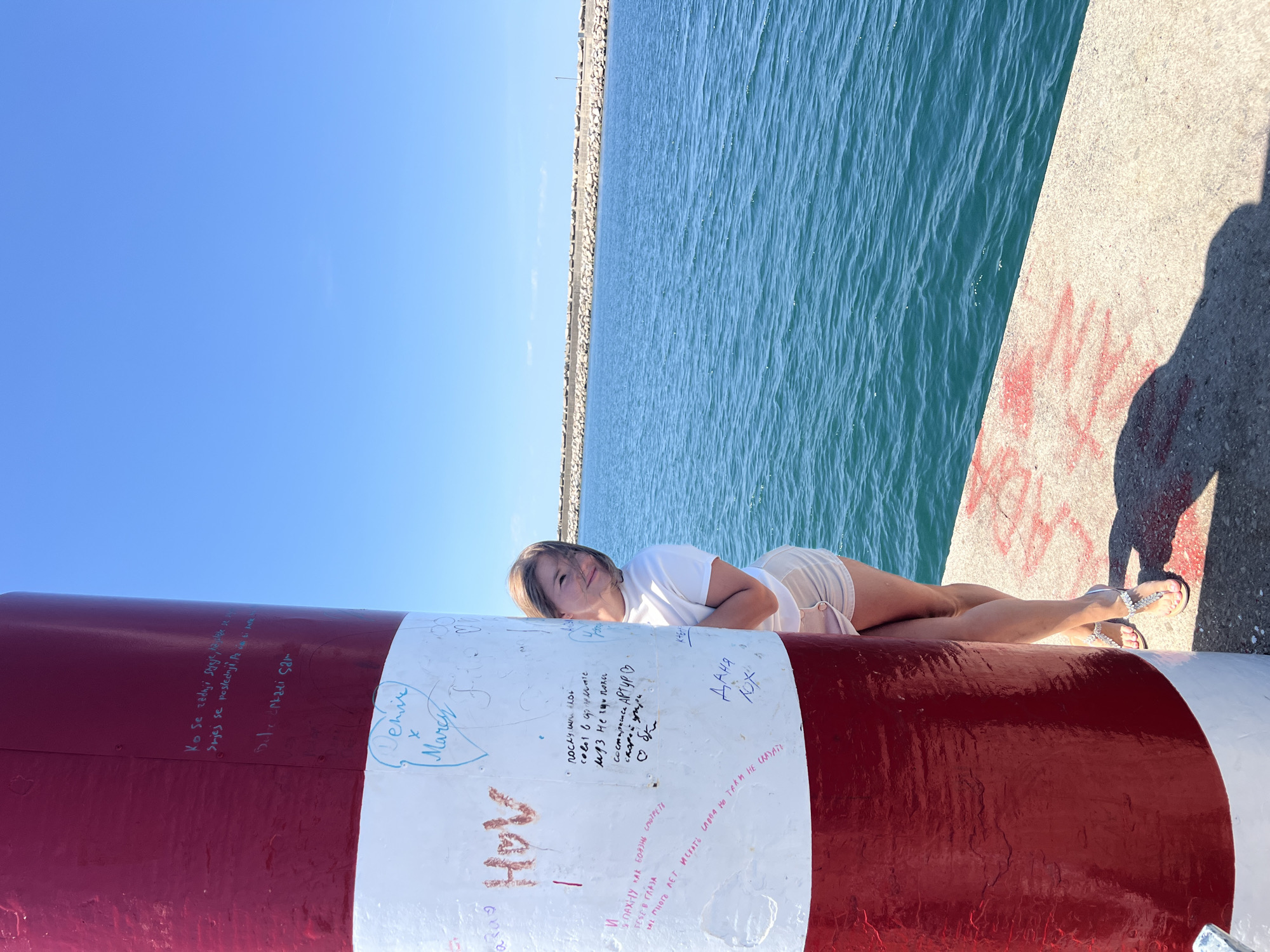 This outdoor, full-length image depicts a young woman posing on a large, graffiti-covered red and white striped navigational pillar or buoy on a sunny day in Bar, Montenegro. The setting features a clear blue sky and calm, vibrant blue-green sea water, with a concrete pier or breakwater visible along the horizon.
The woman, with light brown hair, is dressed in a white t-shirt, light-colored shorts, and embellished white sandals. She lies comfortably on her side on the pillar, resting her head on her right arm, and looks towards the camera with a relaxed, gentle smile. Her left arm is bent, with her hand resting near her stomach.
The large red and white pillar in the foreground is adorned with numerous handwritten messages and signatures in various colors and languages, predominantly Cyrillic and Latin script. Notable visible text includes "Denise x Marco" within a drawn heart, "Даня NOX", and "Артур В". Phrases such as "Ko se zadnji styeje, slede se Srjes se poslednji" (roughly: "Whoever comes last, waits last") and "КАК БОЯЗНЬ СМОТРЕТЬ ТЕБЕ В ГЛАЗА" (How scary it is to look into your eyes) are also present. The concrete surface of the pier shows additional red graffiti and a distinct shadow, likely cast by the person taking the photograph. The overall scene evokes a relaxed, summery ambiance by the sea.