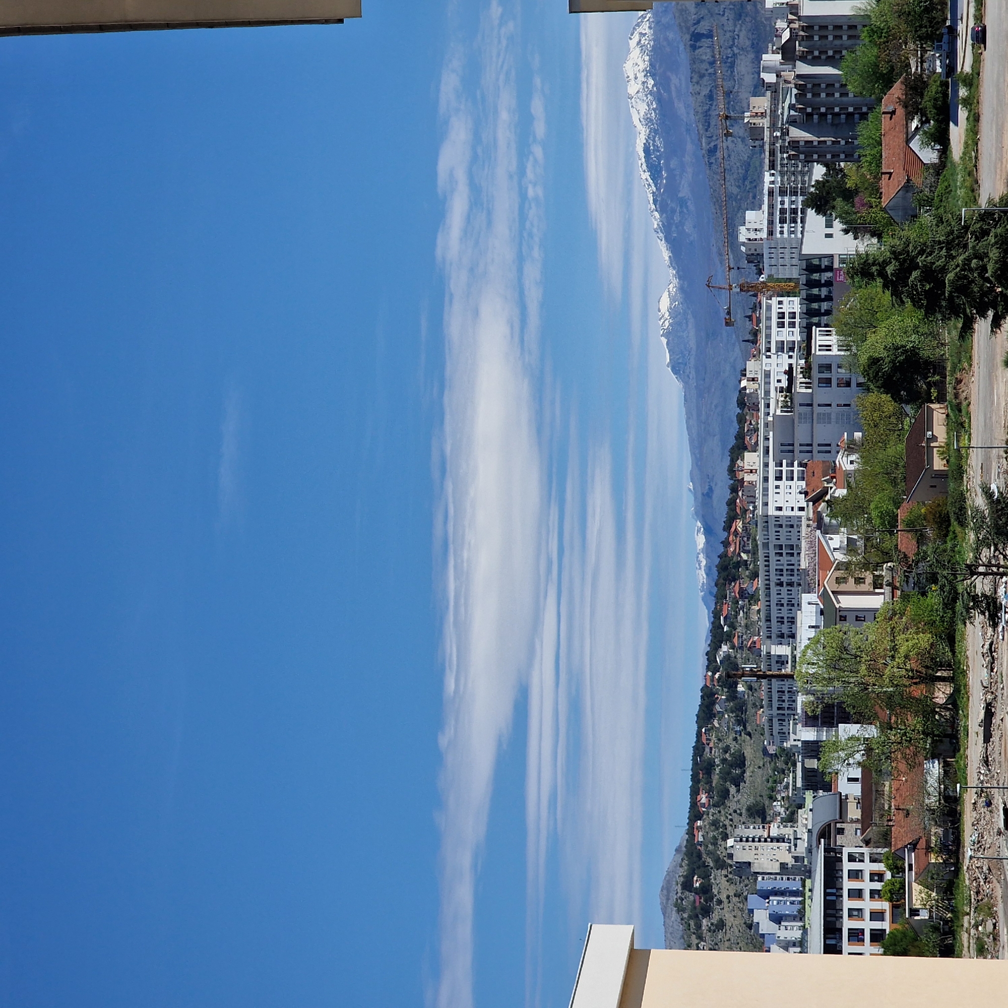The image captures a scene in Podgorica, Montenegro, featuring a cityscape juxtaposed against a mountainous backdrop. The sky is predominantly blue with streaks of white clouds vertically oriented. Buildings of various heights and architectural styles, largely white and modern, dominate the midground, interspersed with trees and residential houses with red-tiled roofs. A yellow construction crane is visible among the buildings. The background features a mountain range with snow-capped peaks. A roadside with trees and debris is present in the foreground.
