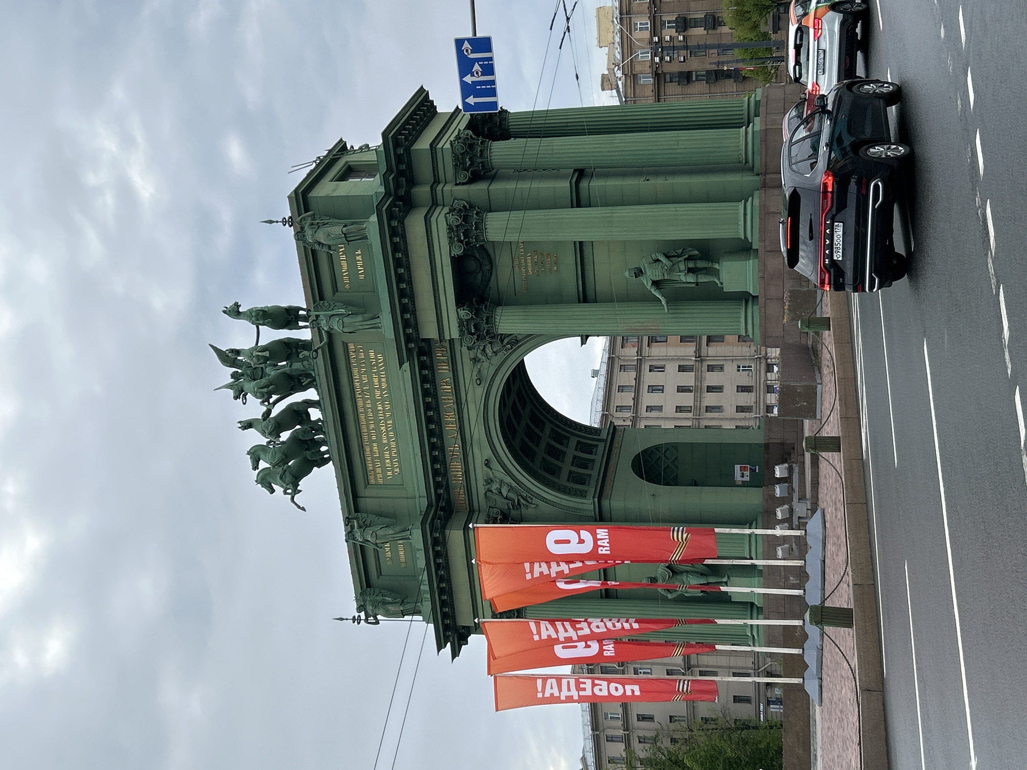 The image shows the Narva Triumphal Arch in Saint Petersburg, Russia. The arch is a large, green structure with columns, statues, and inscriptions. There are several orange flags with white text. Cars are driving on the street next to the arch. The sky is overcast.