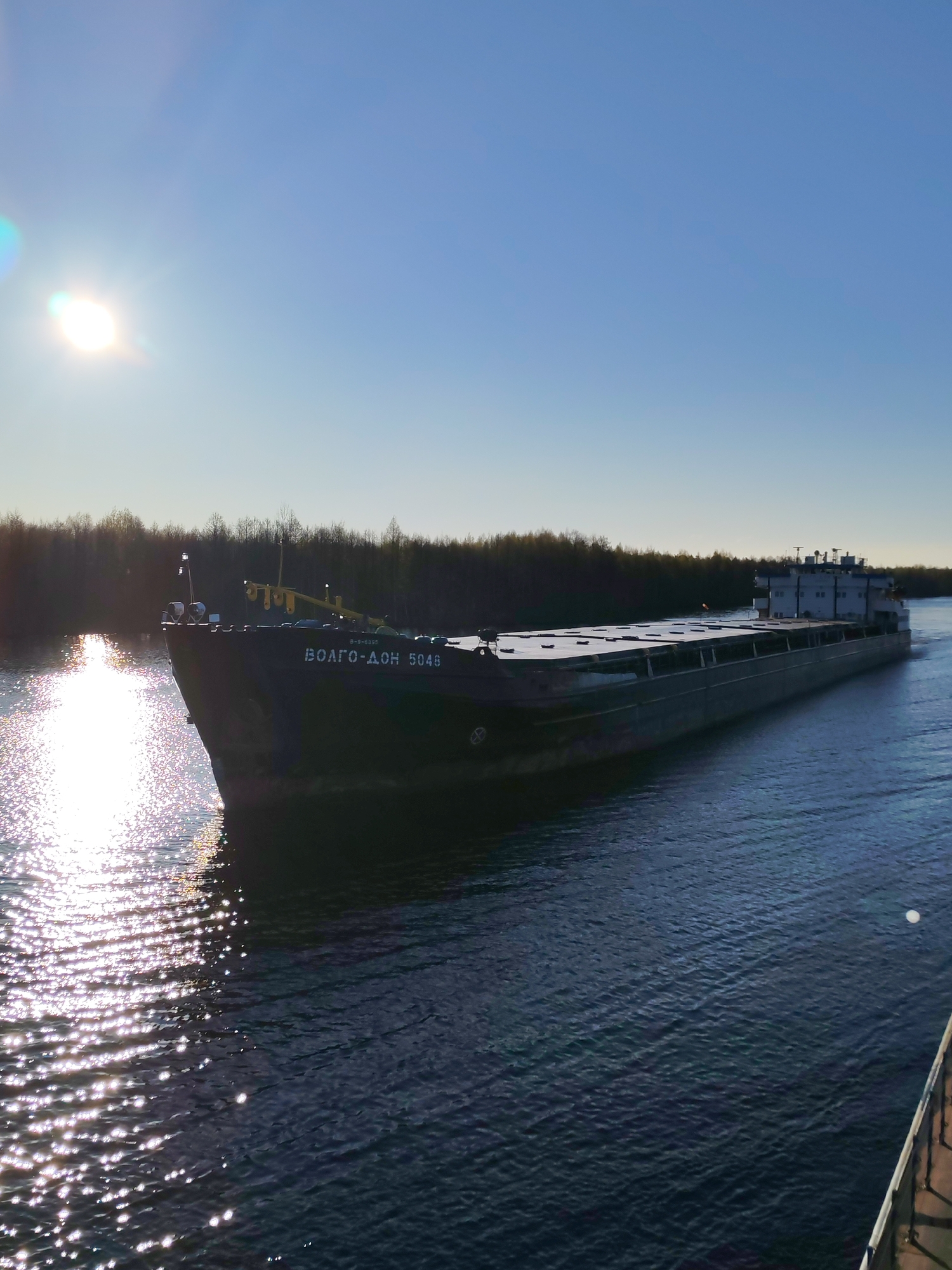 A cargo ship, marked "ВОЛГО-ДОН 5048," is moving through a body of water, possibly a river or canal. The sun reflects off the water in front of the ship, and trees line the distant bank. The image appears to have been taken in Novokemskii, Russia.