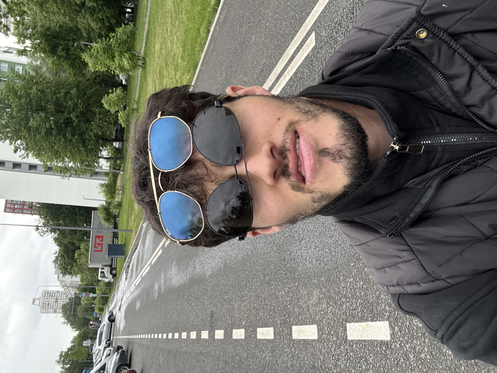 The image shows a man standing on a road in Moscow, Russia. The road appears to be wet, and there are white line markings along its length. Behind the man and road, there is a row of parked cars, followed by green grass and trees, with tall buildings in the distance. The man is wearing a black jacket, and two pairs of sunglasses, one stacked on top of the other. The top pair has black lenses, while the bottom pair has blue lenses. He has a beard and is looking at the camera with his mouth slightly open.
