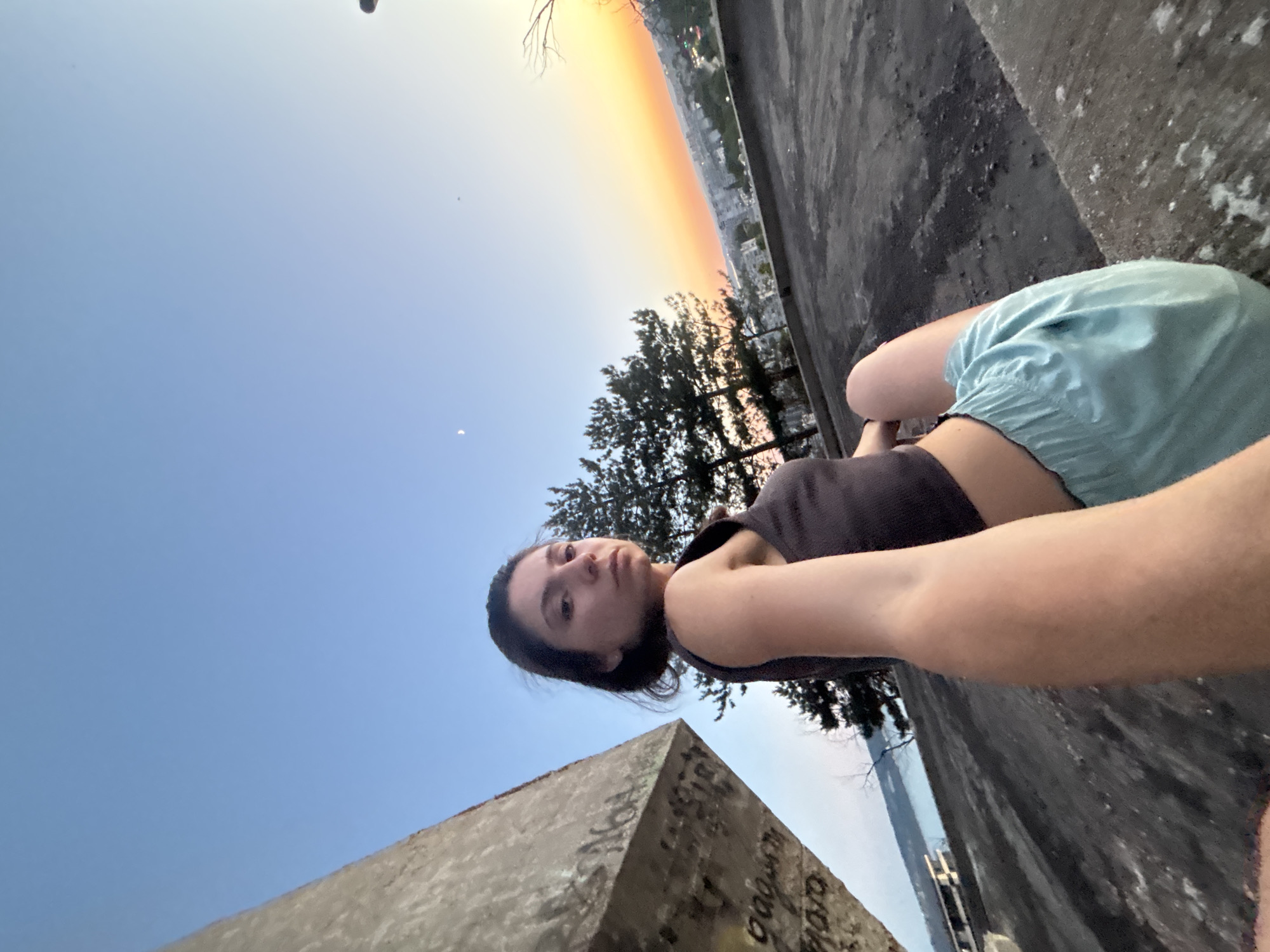 In Varna, Bulgaria, at sunset, a young woman sits on a concrete structure with graffiti. She is wearing a dark grey tank top and light blue shorts. Behind her, pine trees and a distant city skyline are visible, illuminated by the warm glow of the setting sun. A sliver of the moon is present in the clear, pale blue sky. The foreground is dominated by the rough texture of the concrete, with hints of scattered debris. A body of water can be seen in the far left of the image.