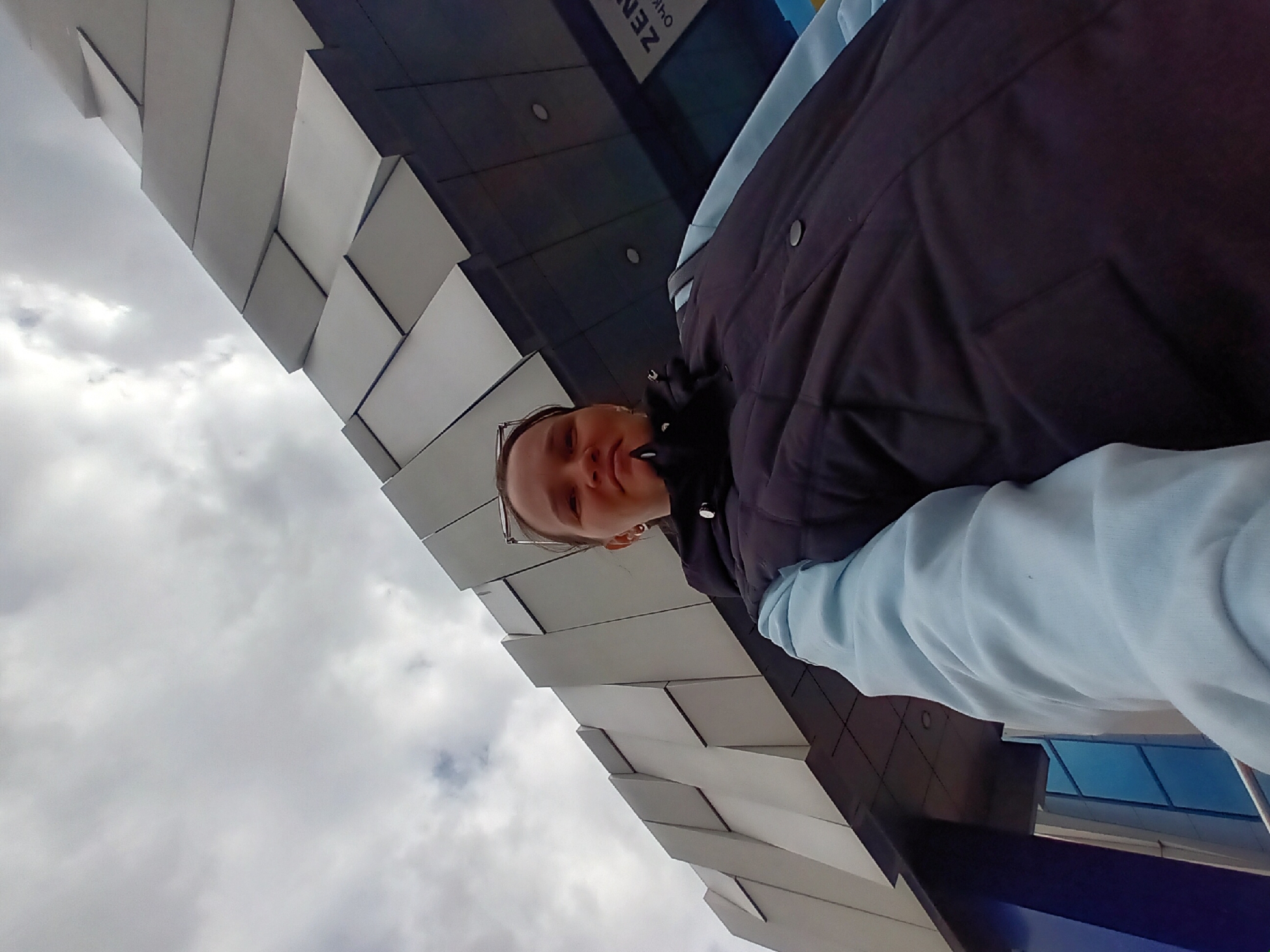 The original image is a vertically oriented selfie, rotated 90 degrees counter-clockwise. When viewed upright, it captures a young woman looking at the camera from a low angle, standing in front of a modern building under a cloudy sky in Chaikovsky, Russia.
The woman, positioned in the lower left portion of the frame, appears to be in her 20s or 30s. She has dark hair pulled back, wears glasses on her head, and displays a calm expression with a slight smile. She is dressed in a dark, possibly black or navy, puffer vest layered over a light blue long-sleeved top or sweatshirt.
The background features a distinctive building facade, diagonally cutting across the frame. This facade is characterized by numerous light-colored, geometric, cube-like panels arranged in a staggered, three-dimensional pattern, creating a modern, textured surface. Behind these panels, parts of the building appear dark. On a blue section of the building, blurry Cyrillic text is discernible, potentially reading "ZEN" (ЗЕН) and "OKNO" (ОКНО).
The upper right section of the image is filled with an overcast, cloudy sky, indicating daytime, likely on a cool day. The low-angle perspective looks upwards at both the woman and the building, highlighting the architectural design. The image captures a casual outdoor moment.