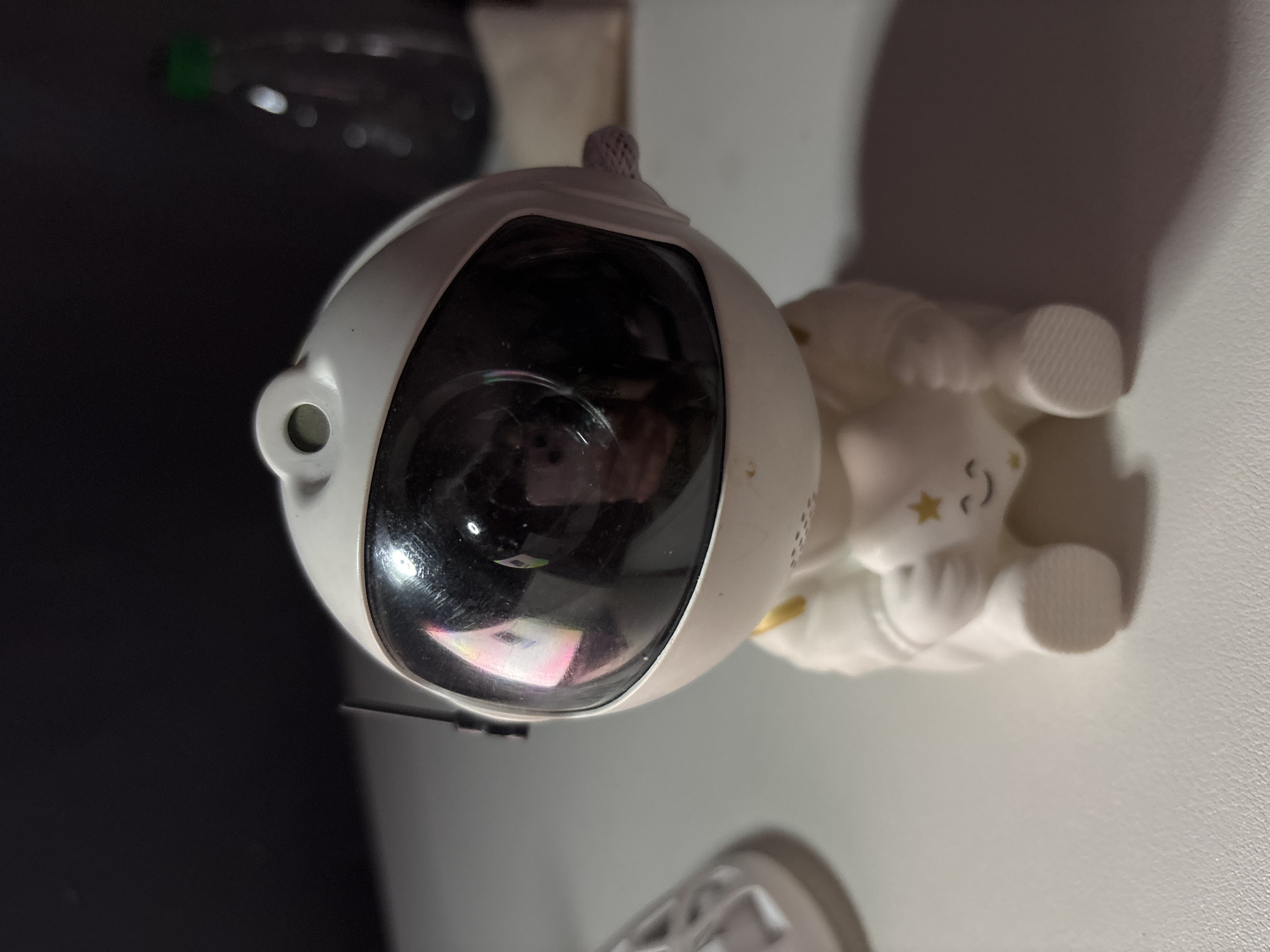 An astronaut figurine is displayed on a white surface, with a dark background behind it. The figurine is white with gold accents and is holding a star. The figurine's helmet visor reflects light. In the background, a blurred bottle with a green cap is visible. The image appears to have been taken indoors. There is no indication of location in Pastavy, Belarus.