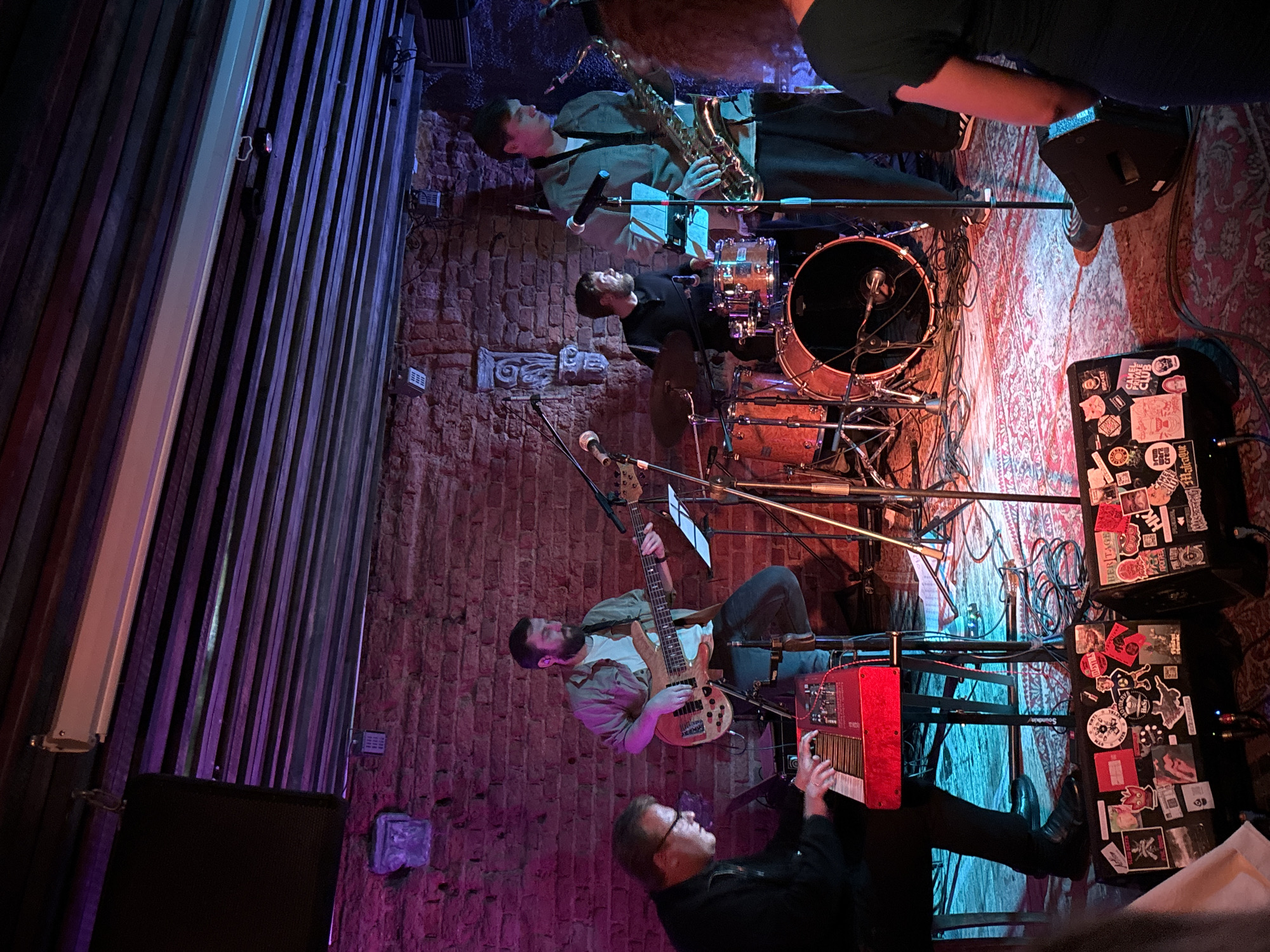 The image shows a band performing live on a stage in what appears to be a small venue. The background consists of a brick wall. The lighting is dim and atmospheric, with purple and blue hues dominating the scene. In the foreground, a keyboard player with glasses is seated on the left, playing a red keyboard. To his right, a man with a beard is playing a bass guitar. Behind them, a drummer is seated at a drum kit. On the far right, a man is playing a saxophone. There are microphones and music stands set up on the stage. The floor is covered with a patterned rug, and there are speakers placed in front of the stage, adorned with stickers. The overall impression is that of a lively, intimate concert. The location is identified as a club in Minsk, Belarus.