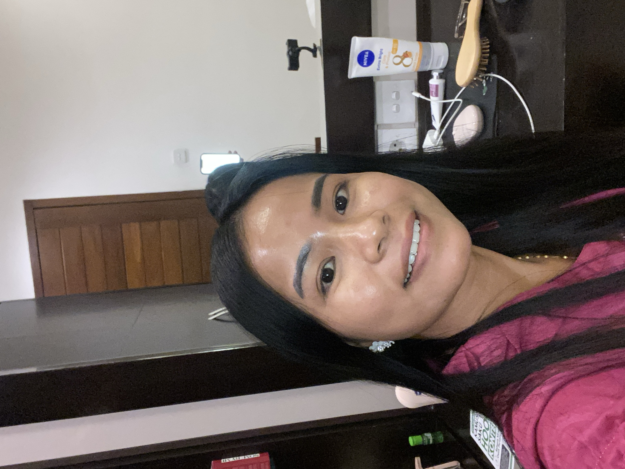 A selfie taken in what appears to be a bathroom setting in North Kuta, Indonesia. The subject is a smiling woman with dark hair and a red shirt. Behind her are bathroom fixtures including a counter, a sink, a wooden door, and various cosmetic items such as Nivea lotion and makeup.