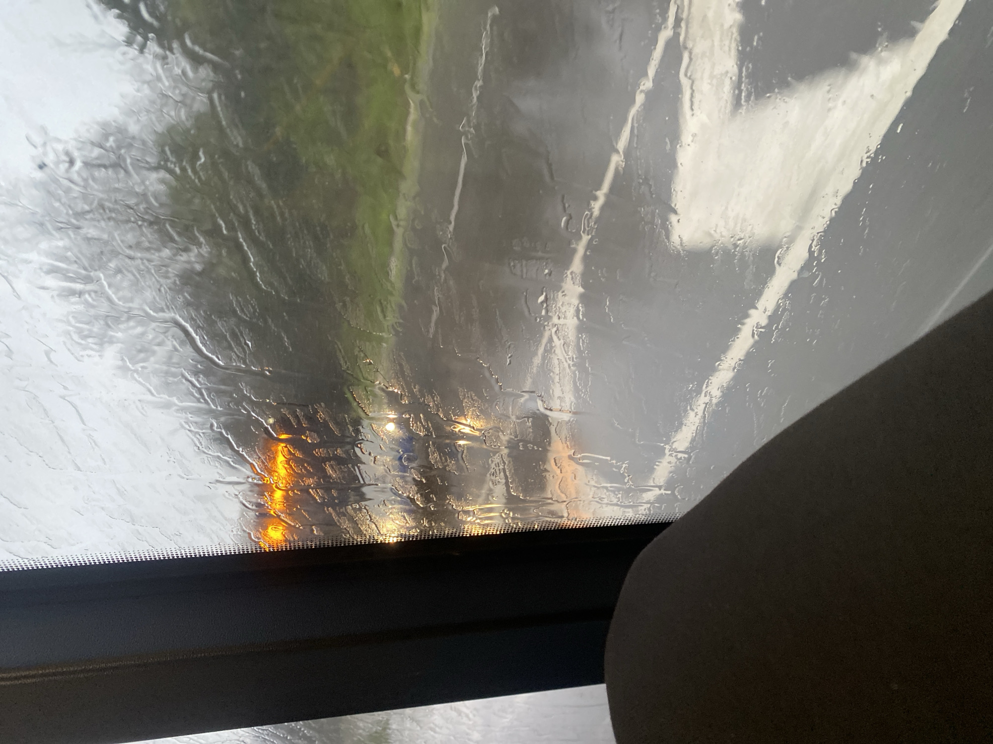 The image depicts a view through a rain-streaked windshield, likely inside a vehicle. The wiper line is visible. Outside, there is the impression of a road with white lane markings and green vegetation. Headlights are visible in the distance. The location is unspecified, and there is no evidence indicating Varna, Bulgaria.