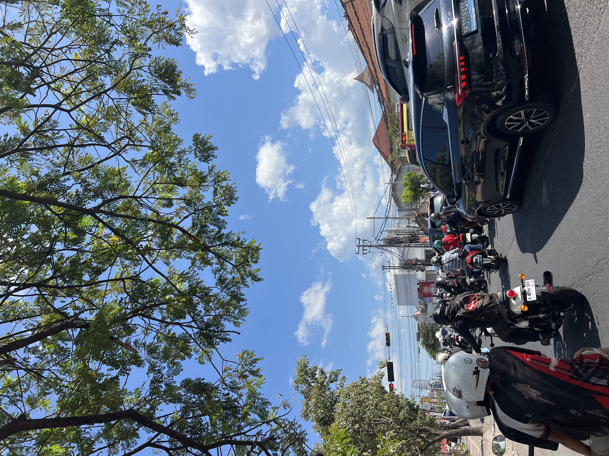 The image depicts a busy street scene in Denpasar, Indonesia, on a sunny day. The sky is a vibrant blue, dotted with fluffy white clouds. Lush green tree branches hang over the scene from the top left of the frame, casting dappled shadows. A traffic light shows red, indicating a halt for the vehicles.
The road is filled with motorcycles and a few cars. Several individuals on motorcycles, wearing helmets for safety, are either stopped or moving slowly in traffic. One person in the foreground is wearing a white helmet and a black, red, and white patterned shirt. Another person on a motorcycle in front of them wears a black helmet and a backpack. A black SUV is prominently featured in the mid-right of the image, with its license plate visible. Buildings line the street, with tiled roofs characteristic of Indonesian architecture. Power lines and poles are visible overhead, typical of urban infrastructure. The overall impression is one of a typical, bustling day in the city.