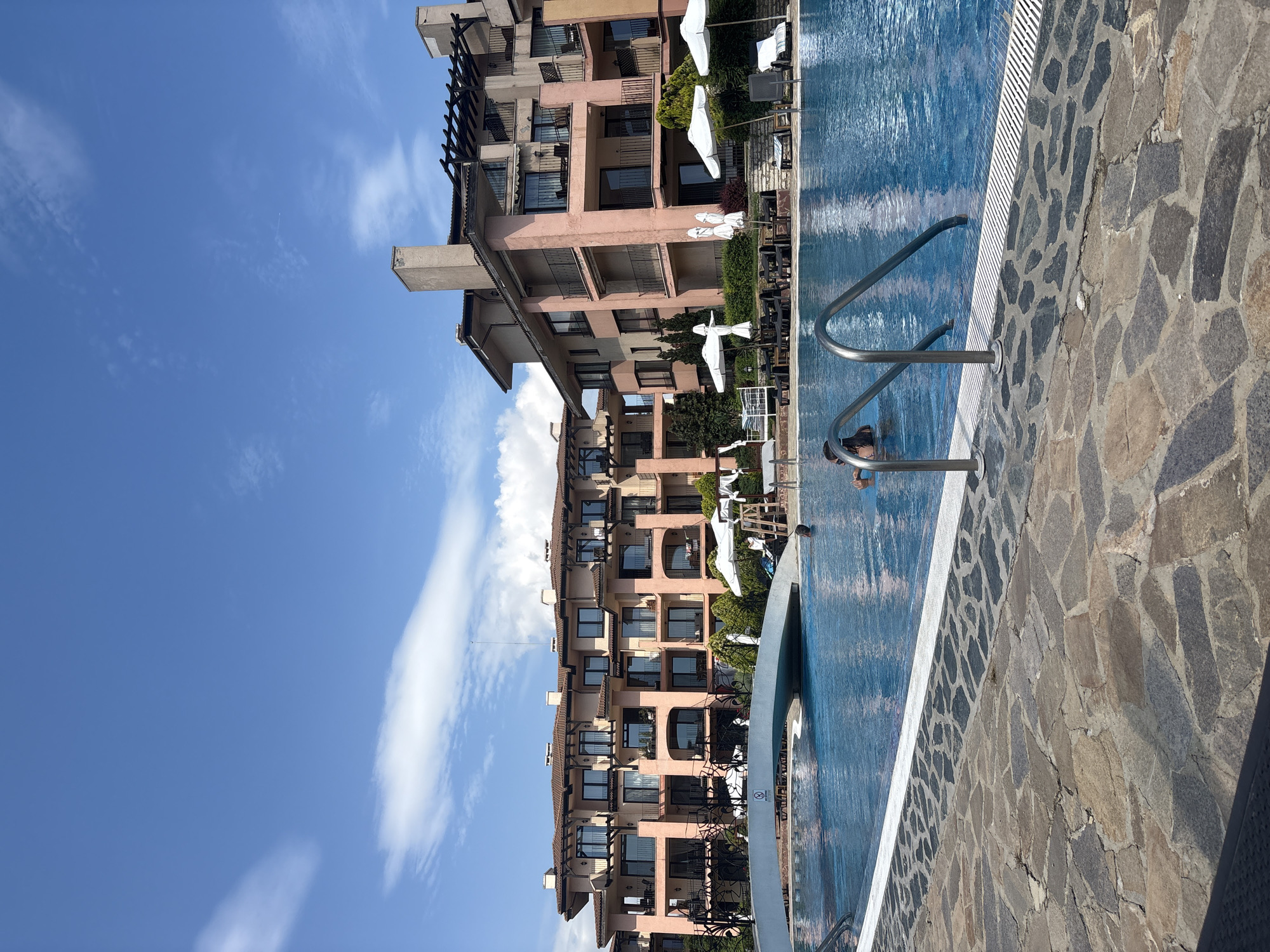 A resort pool area is depicted with a large, blue swimming pool in the foreground. The pool features a stone patio surrounding it, with metal railings leading into the water. In the background, a multi-story building with a pink facade and balconies can be seen. Several white umbrellas are set up by the pool, along with lounge chairs and tables. One person is partially visible swimming in the pool, and another person is sitting on a lounge chair. The sky is clear blue with scattered white clouds.