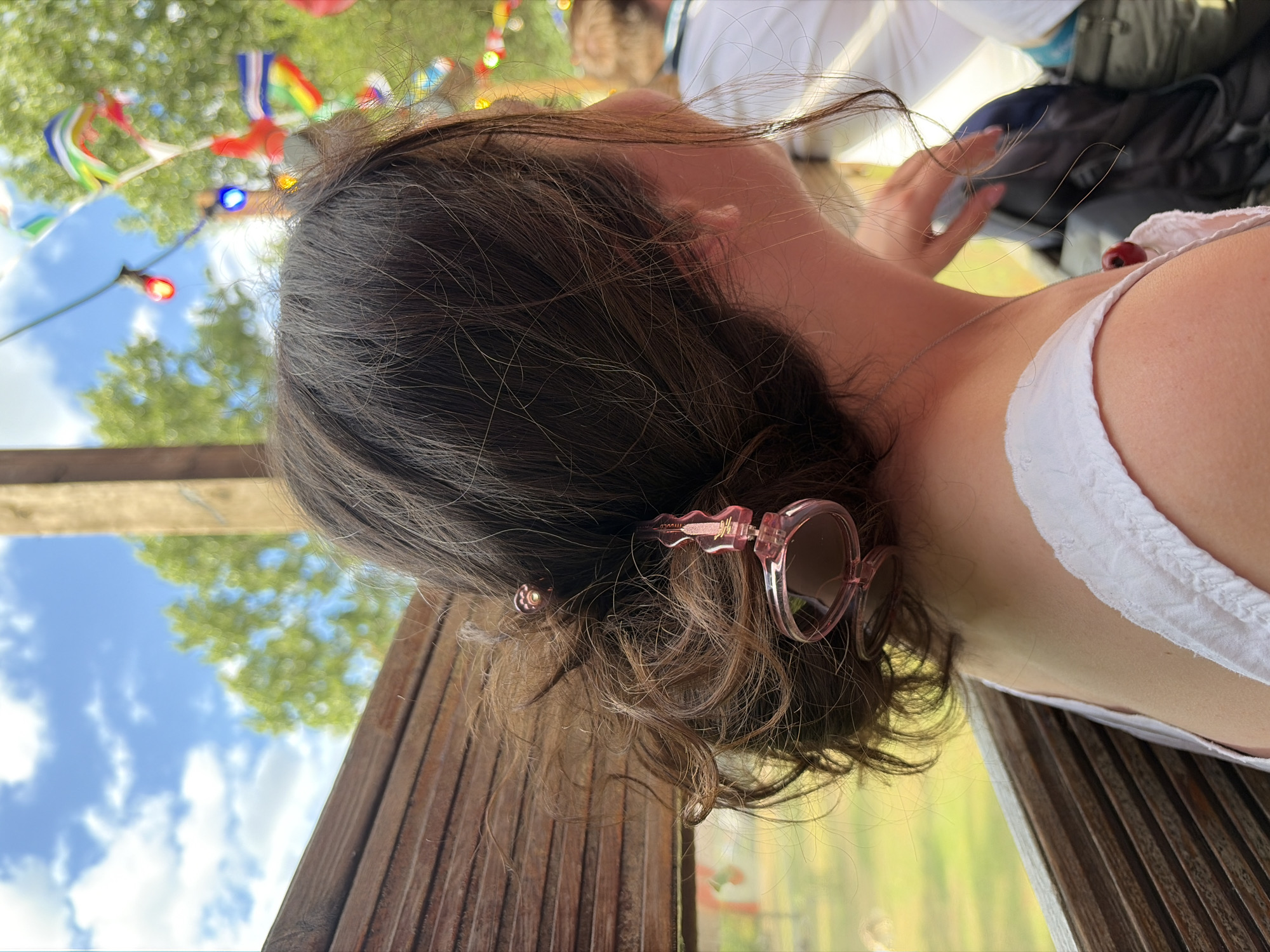 The image shows a close-up, sideways view of a person with dark, wavy hair. Pink-tinted sunglasses are perched on the back of their head, and a small silver pendant rests on their collarbone, near a red cherry that is resting on their white top. The person is outdoors, with a backdrop of trees, a bright blue sky with scattered clouds, and a string of colorful flags or pennants. A wooden structure, possibly a fence or seating, is visible in the foreground. The lighting suggests it is daytime, with clear weather. The overall impression is one of a casual, outdoor gathering or event.