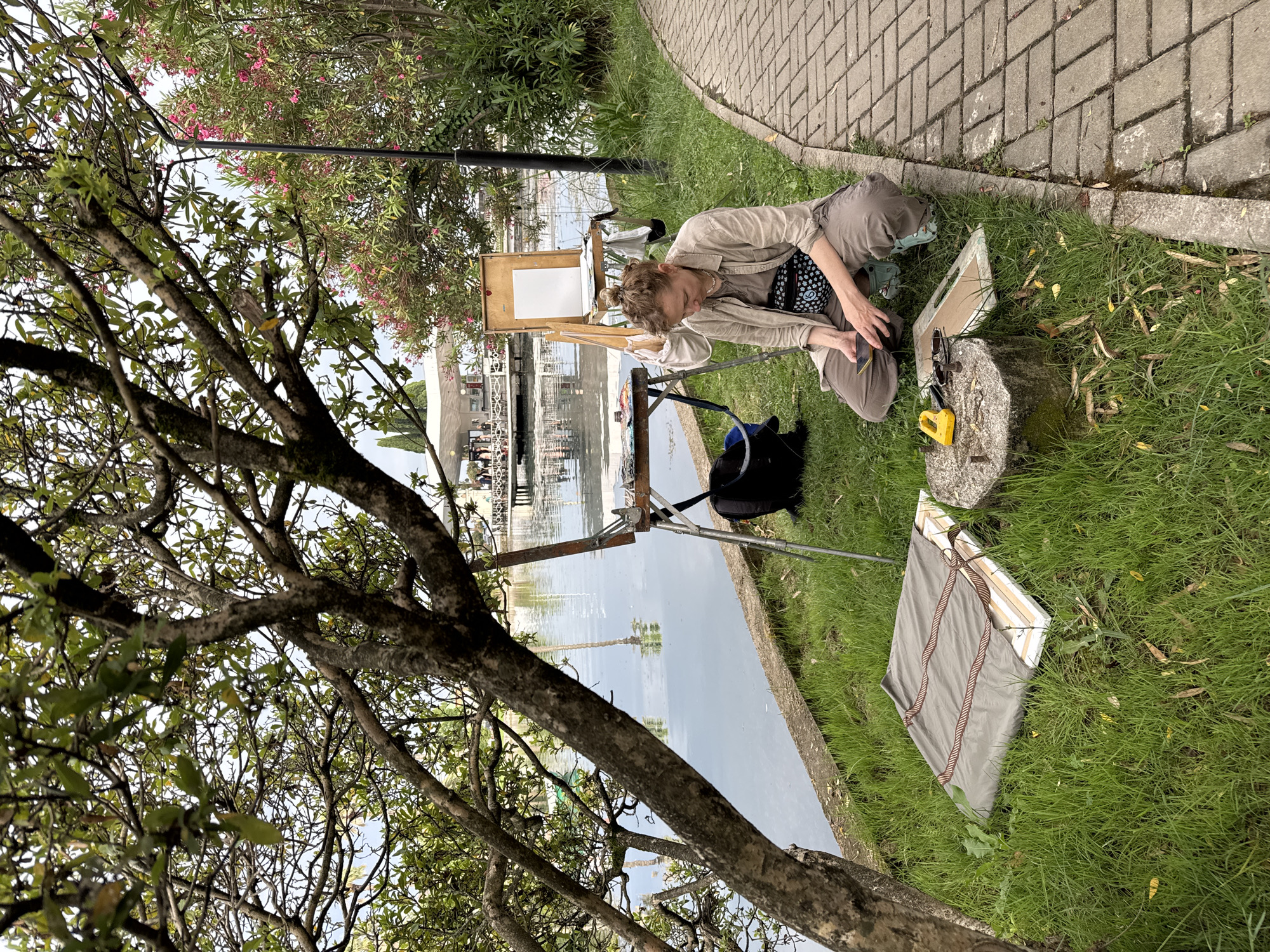 In New Athos, Abkhazia, on a cloudy day, a woman is sitting on the grass next to a paved walkway, by a body of water. She is wearing a light brown button-down shirt, grey pants, and light blue clogs, with a black fanny pack around her waist. She is holding a smartphone in her hands and appears to be looking at it.
In front of her on the grass is a concrete block with a yellow tool and some other objects. Next to the concrete block, also on the grass, is a canvas covered with a grey tarp and tied with rope.
Behind the woman, an easel holds a blank white canvas. To the left of the easel is a table with art supplies, including tubes of paint. A black backpack is placed on the grass near the table.
The background shows a calm body of water with reflections, and on the opposite side of the water, there is a white building with a pier and some people. Green trees and pink flowers are visible in the surrounding vegetation. The ground is covered with grass and a paved walkway.