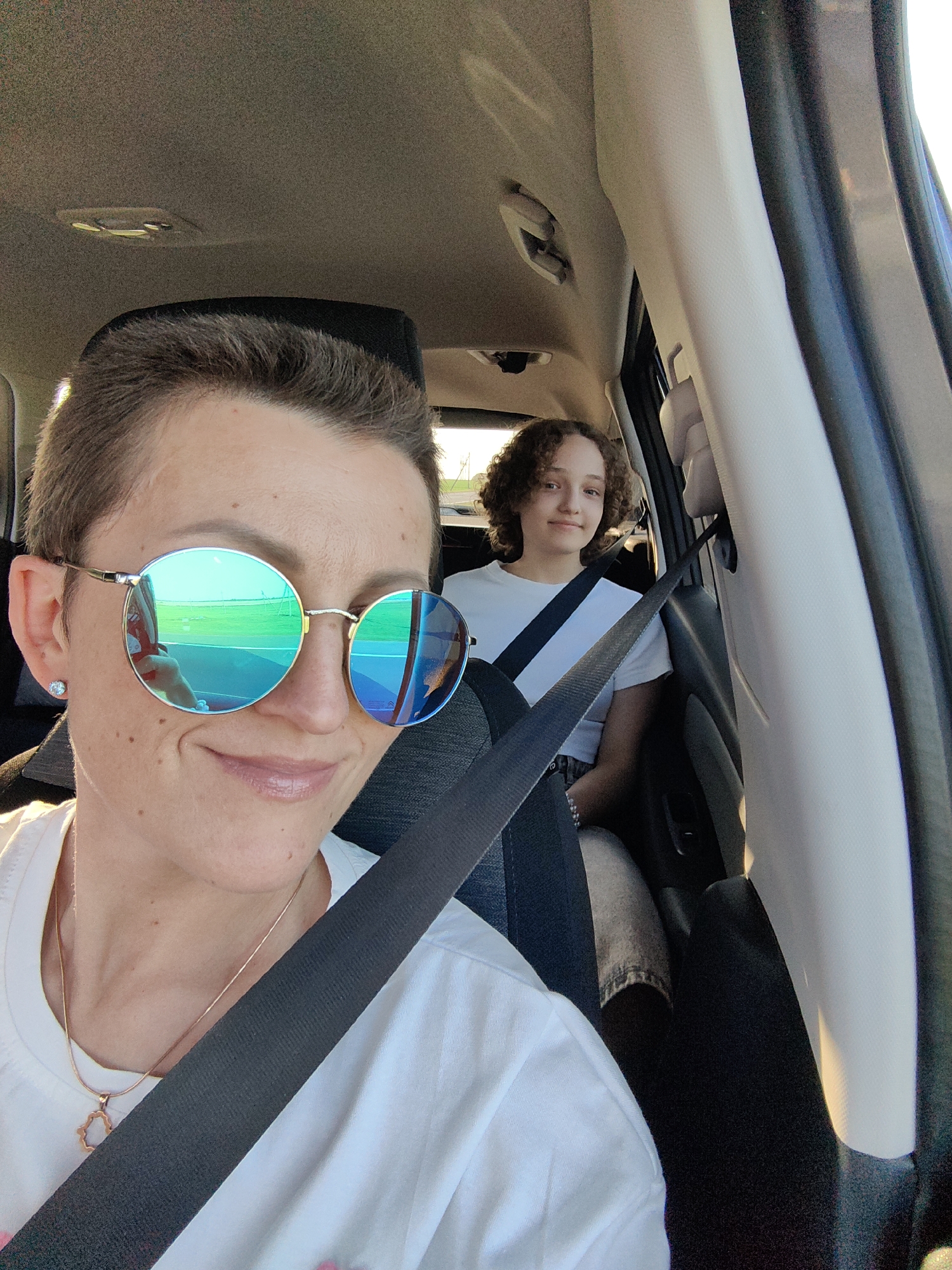 In the foreground, a person with short brown hair and round, mirrored sunglasses smiles at the camera. They are wearing a white t-shirt, a thin rose gold necklace with a pendant, and a seatbelt. In the background, a younger person with curly brown hair and a white t-shirt is seated. They are also wearing a seatbelt. The image appears to be taken inside a vehicle, possibly in or around Міханавіцкі сельскі Савет, Belarus. The lighting suggests it is daytime.