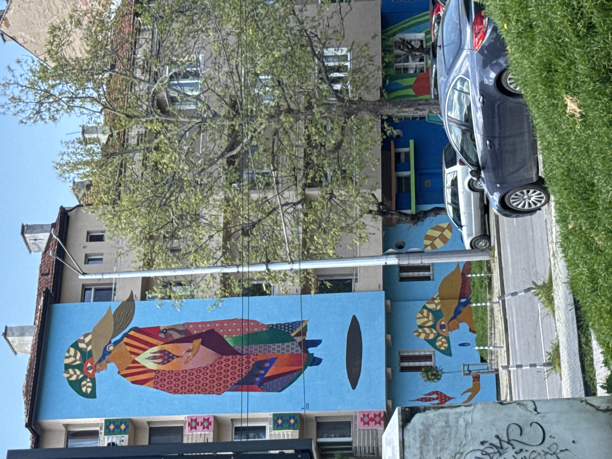 The image shows a building in Unknown, Unknown, with a large mural painted on its side. The mural features colorful, geometric shapes that form an abstract representation of a figure. The building itself is multi-story, with windows and architectural details visible beneath and around the mural. A tree with green leaves is positioned in front of the building, partially obscuring the mural. A parked car is visible on the street adjacent to the building, and greenery is present along the sidewalk. The sky is blue, suggesting a sunny day.