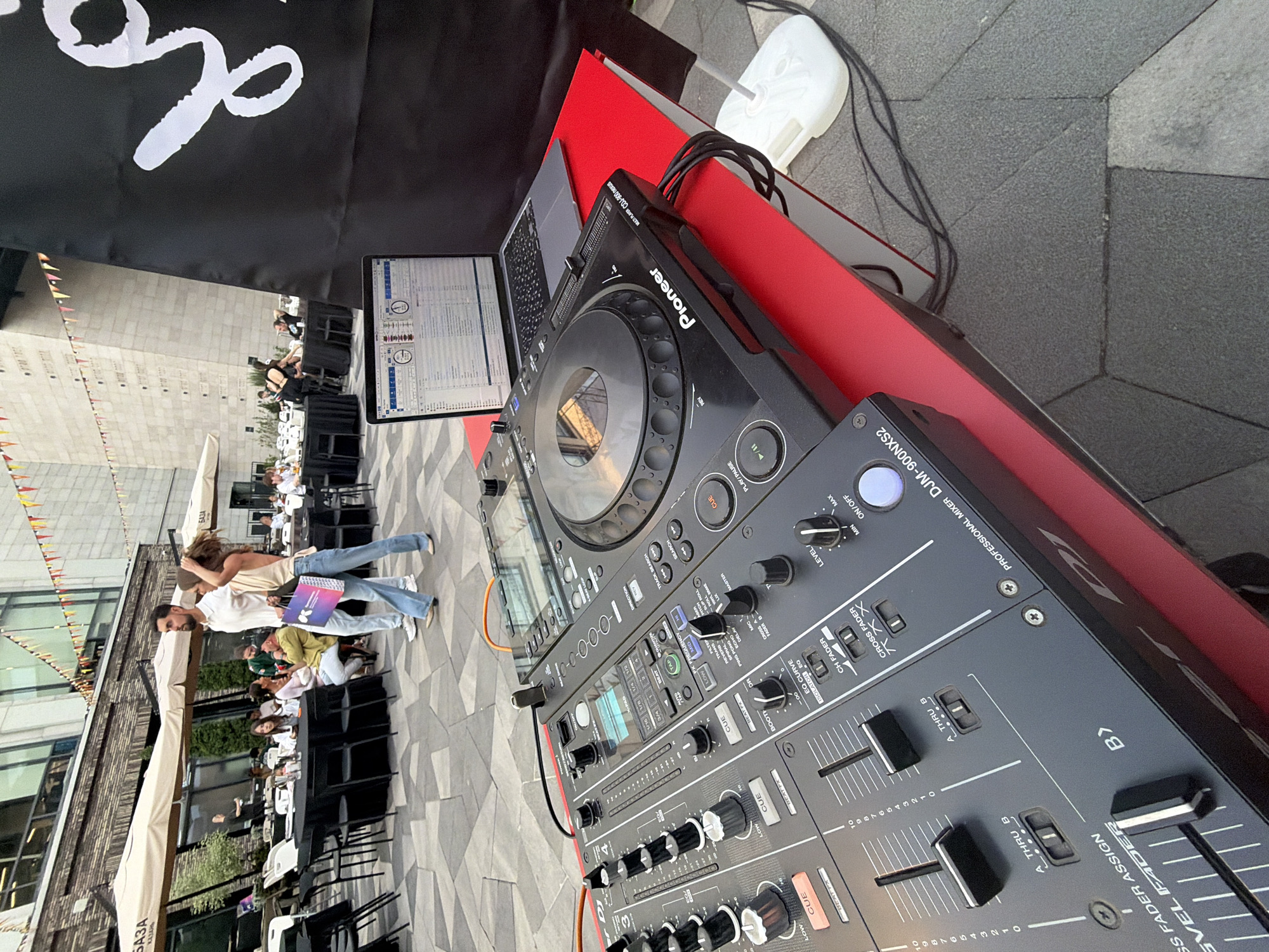 The image shows a DJ setup in what appears to be an outdoor event space, with a crowd visible in the background. The DJ equipment includes a Pioneer DJM-900NXS2 professional mixer and a CDJ-900 Nexus multiplayer. A laptop is open next to the equipment.
The setting is an outdoor area with tables and chairs, suggesting a cafe or restaurant. Based on a sign visible in the background with the text "БАЗА КАЗАНЬ", the location is Kazan, Russia. People are seated at the tables, and two individuals are walking past in the foreground. The surface is paved with patterned stone.