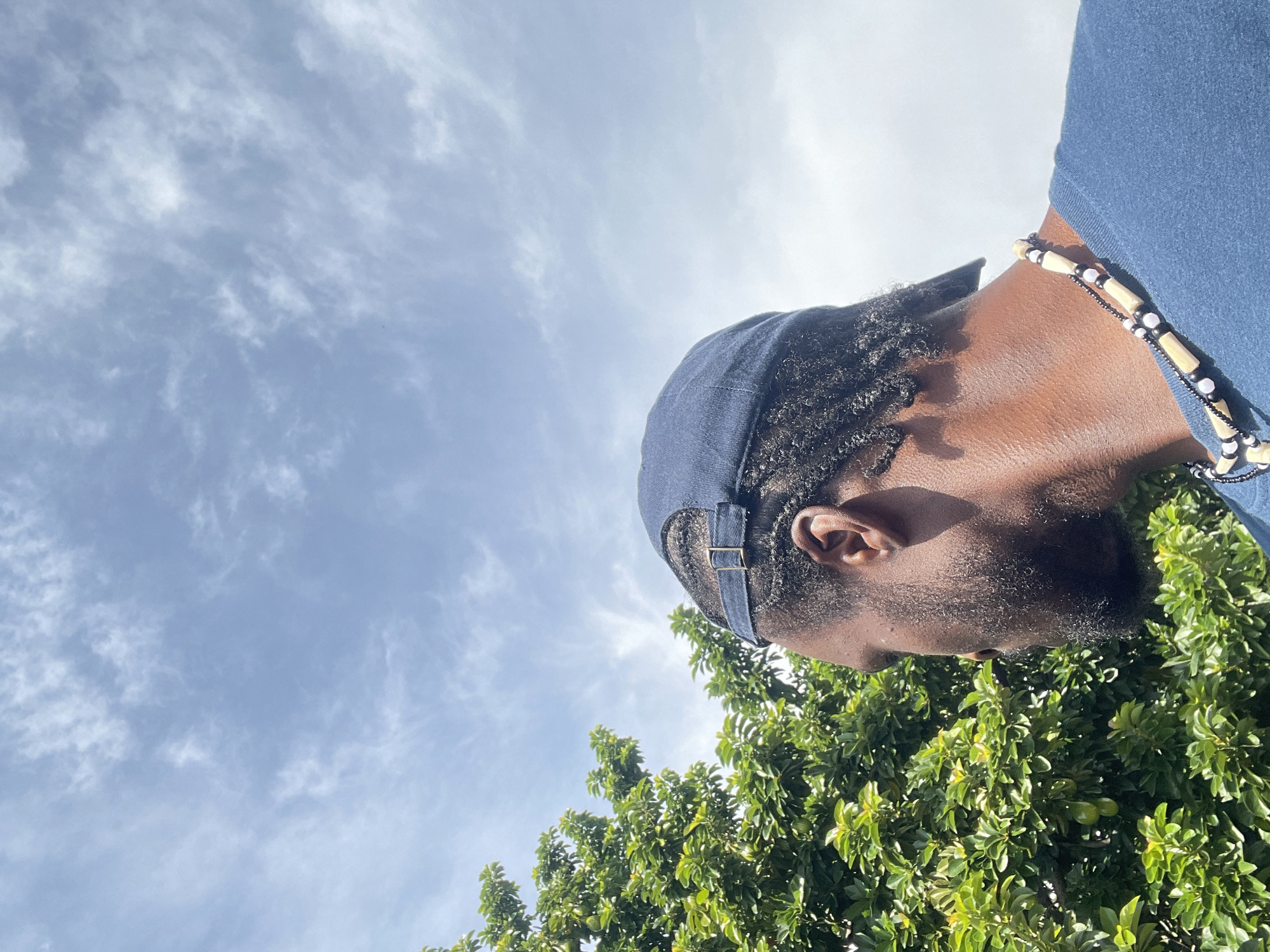 This is a close-up, eye-level, outdoor, medium shot photograph of a Black man wearing a dark blue baseball cap and a dark blue t-shirt. He is also wearing a necklace made of wooden beads and black beads. The man's head is turned to the left, and he is looking away from the camera. His hair is styled in dreadlocks. Behind him, to the left, is a lush green tree with visible avocados. The background consists of a bright blue sky with scattered white clouds. The lighting suggests a sunny day, likely midday. The image does not contain any specific landmarks or elements that would allow for precise identification of the location within Jos, Nigeria.