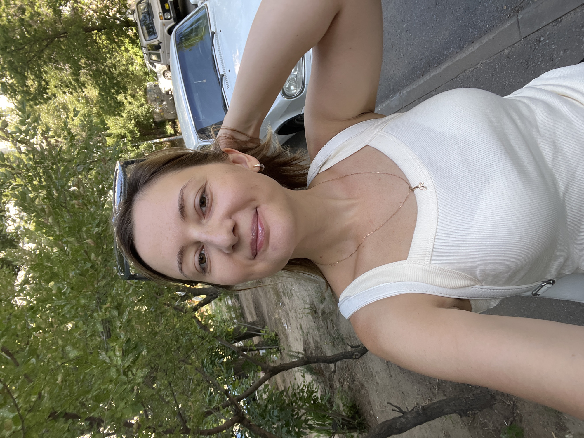 An outdoor, eye-level, medium-shot selfie shows a woman with light skin and brown hair looking into the camera. She has an open-mouth smile and her right hand is behind her head, touching her hair. She is wearing a white tank top and a gold necklace with a small charm. In the background, trees line a dirt path, and two white cars are parked on the street. The scene appears to be a residential street in Volgograd, Russia.
