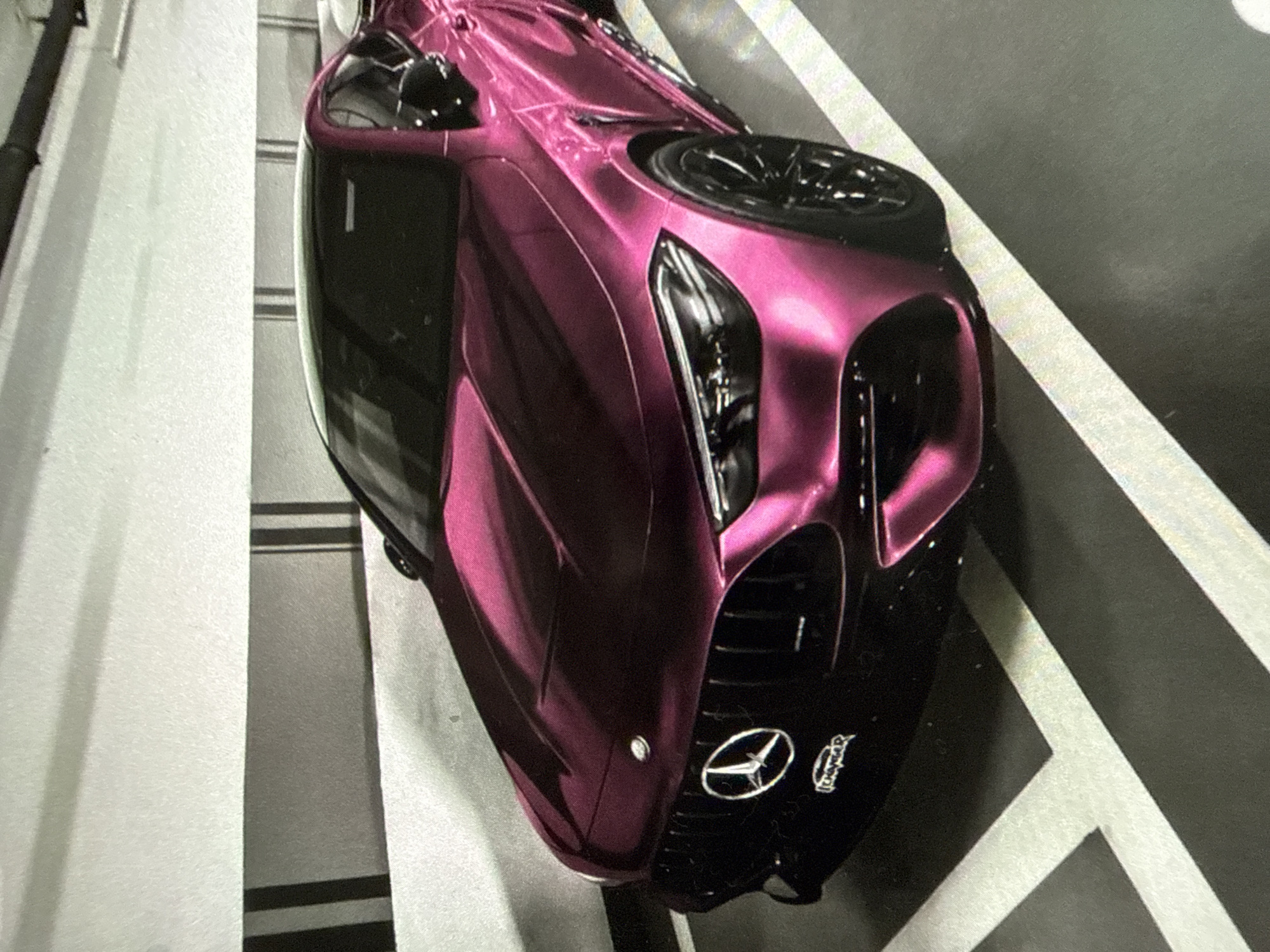 A high-angle shot shows a pink Mercedes-AMG One hypercar parked indoors in an underground parking garage, Unknown. The car is positioned diagonally, with the front right corner nearest the viewer. Visible details of the car include the iconic Mercedes-Benz emblem on the hood, headlights, front grille, and the black roof. The background consists of parking space markings on the floor of the garage. The location is identified as Unknown, Unknown.