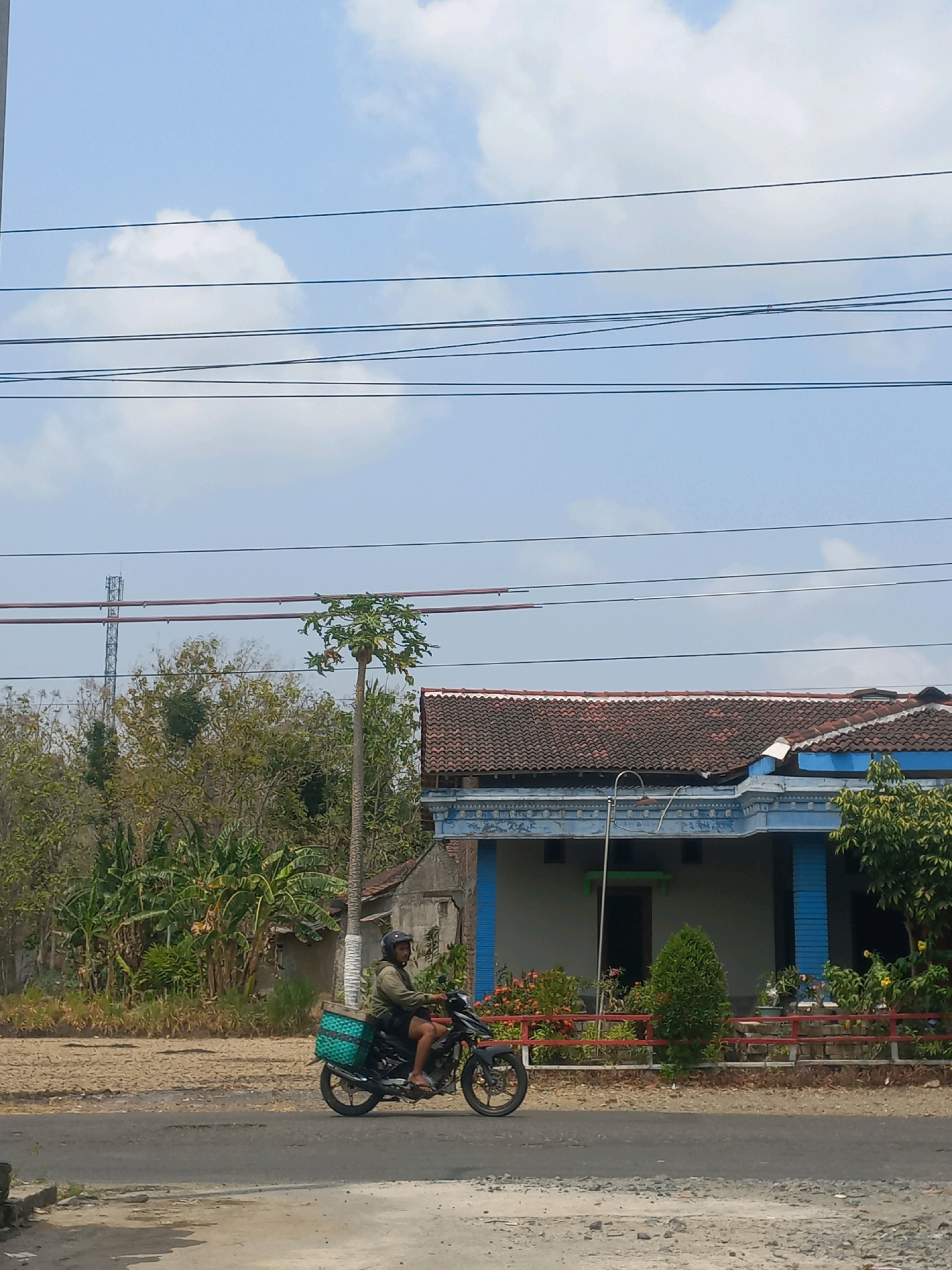 A person rides a motorcycle on a dirt road in Baturetno, Indonesia. The rider is wearing a helmet and a green jacket, and is carrying a green basket on the back of the motorcycle. The motorcycle is black and has a red accent on the handlebars. The road is dry and dusty, and there are trees and bushes on either side. In the background, there is a house with a blue and white facade and a red tile roof. There are also power lines and a cell tower visible in the distance. The sky is blue and there are a few white clouds. The overall scene suggests a rural or semi-rural setting.