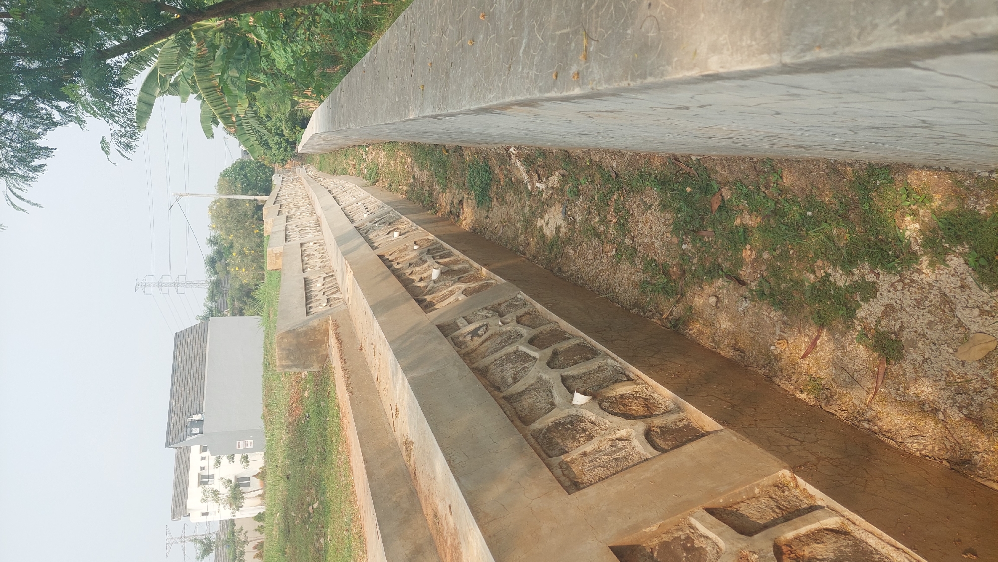 This is a ground-level, eye-level, slightly angled, full shot of a concrete drainage canal in the city of Gunung Putri, Indonesia. The canal walls are constructed with a decorative pattern of interlocking stones. On the left side, a grey, angular building with a dark grey roof and white window frames is visible behind a grassy embankment. Further in the background, a white building with a dark grey roof and multiple windows can be seen. To the right of the canal, a dense cluster of green trees and foliage forms a natural boundary, with banana leaves being particularly prominent. In the far background, a large electricity pylon with visible wires stands against a hazy, light-colored sky. The canal itself appears dry, with sparse patches of green grass and dry earth covering its floor. Sunlight casts shadows, indicating it is daytime.