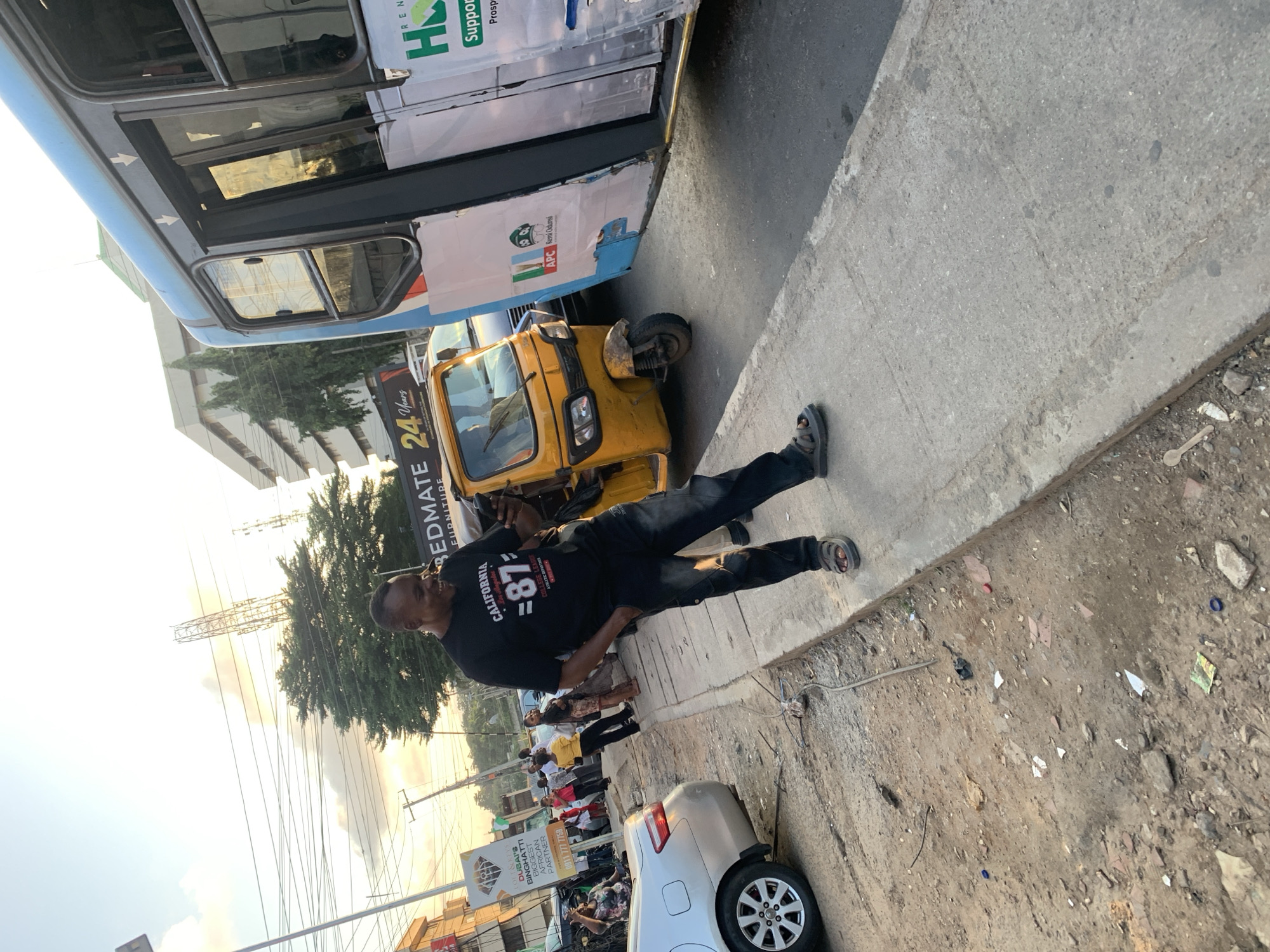 In Ikeja, Nigeria, a Black male is seen standing on a sidewalk, looking at his phone and smiling. He is wearing a black t-shirt with "California 87" printed on it and dark pants. Next to him is a yellow auto-rickshaw, and to his right, a blue and white bus is parked. The environment appears to be an urban street with various buildings and overhead wires visible in the background. The sidewalk is concrete, and the ground beside it is a mix of dirt and debris. Other people and vehicles are present in the background, suggesting a busy street scene. The sky is clear with some clouds, and the lighting suggests it is either early morning or late afternoon.