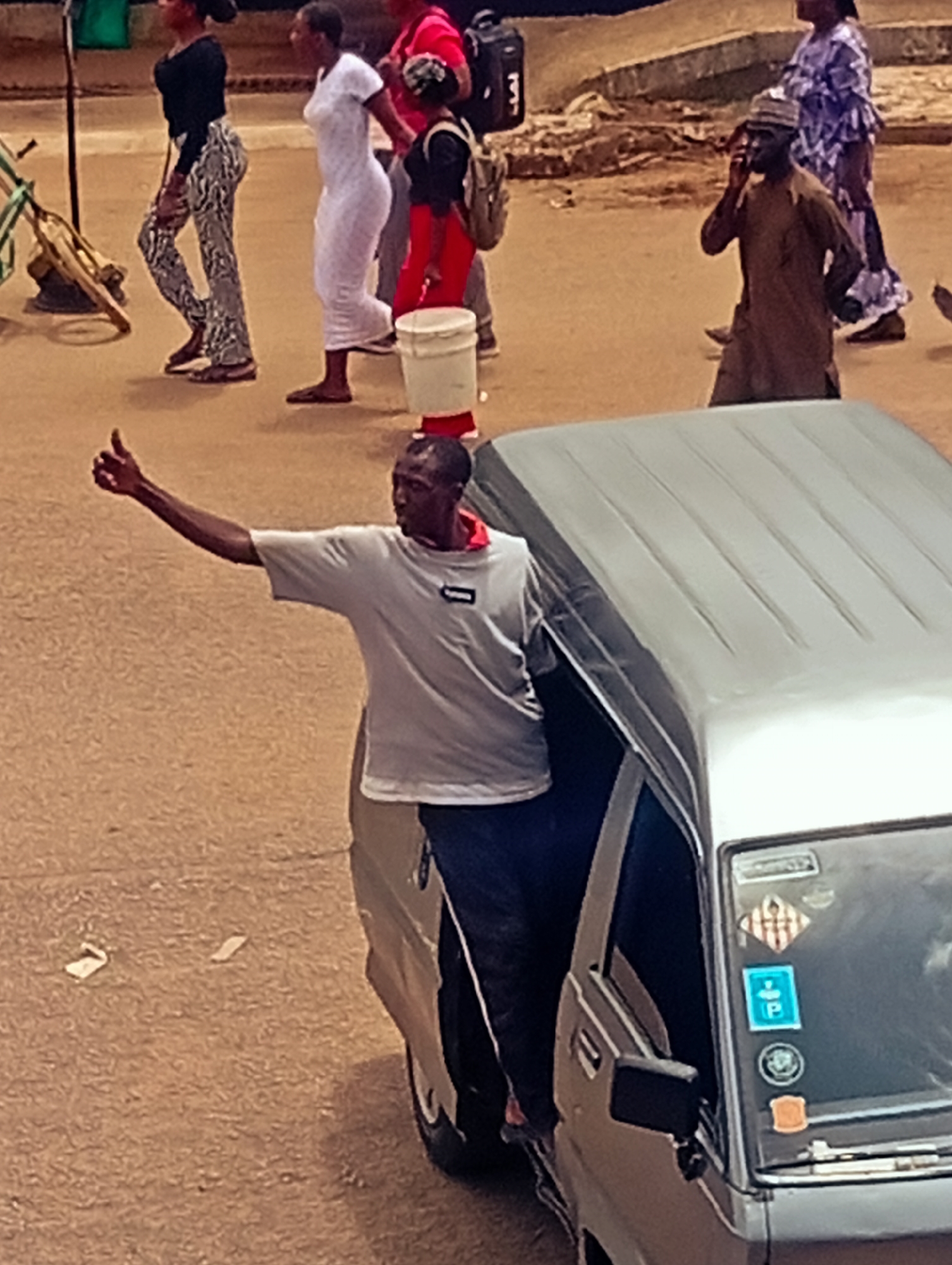 A medium shot, taken from an elevated perspective, shows a man standing out of the passenger side of a parked silver van. The man, who is Black, is wearing a gray t-shirt and dark pants. He has his left arm raised with a thumbs-up gesture. The silver van has several stickers on its front, including a blue parking symbol. In the background, several people are walking on a street. There are 3 women in the back that are walking to the left of the image. In the mid right is a man on his phone wearing a brown cloak and cap. The scene takes place outdoors, likely in the city of Bukuru, Nigeria, as inferred from the metadata. The road is a light, earthy color.