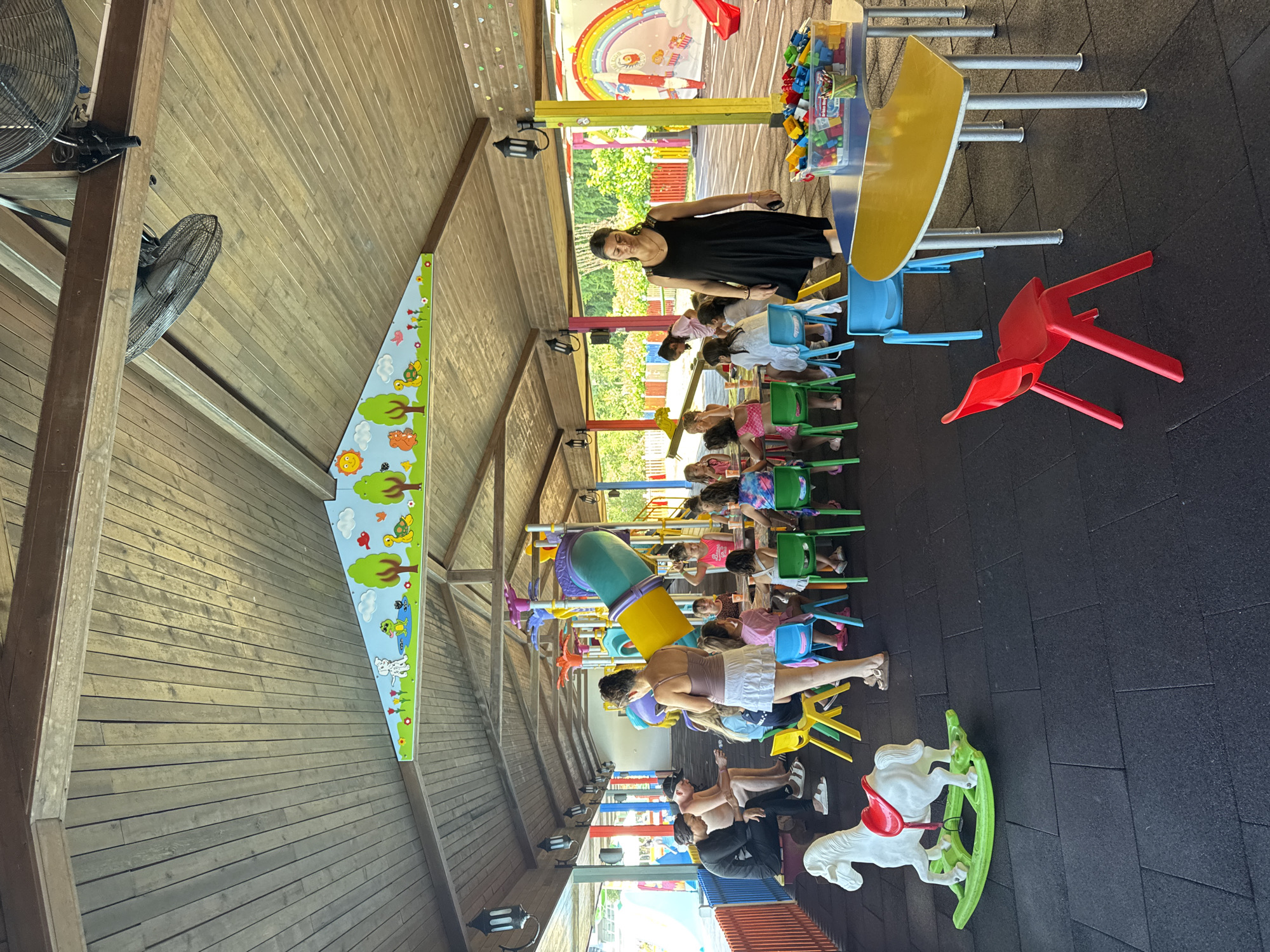 The image depicts a colorful indoor playground or activity area, likely part of a resort or children's facility, located in Aksu, Turkey. The space is covered by a wooden slatted roof supported by wooden beams, with large industrial fans mounted overhead. Decorative, cartoon-like murals featuring animals and nature are visible along the upper walls and the underside of the roof.
In the foreground, a white rocking horse with a red saddle sits on a black rubberized floor. Several small, brightly colored chairs (red, yellow, blue, green) are arranged around small tables. One table has a clear plastic bin filled with colorful building blocks and pencils.
A group of children, primarily girls, are seated at tables, appearing engaged in activities or snacks. Some adults are also present. A woman in a black dress is walking through the scene, holding a phone. To the left, a few adults are seated on a bench, and a woman in a white skirt stands near the children's tables.
In the background, a large, multi-colored playground structure with slides and climbing elements is visible. A prominent mural of a rainbow arching over a rocket ship and clouds adorns a wall on the right side of the scene. Fencing and greenery are visible through open sections of the structure, suggesting an outdoor element or view. The overall atmosphere appears to be a lively and supervised children's entertainment area.