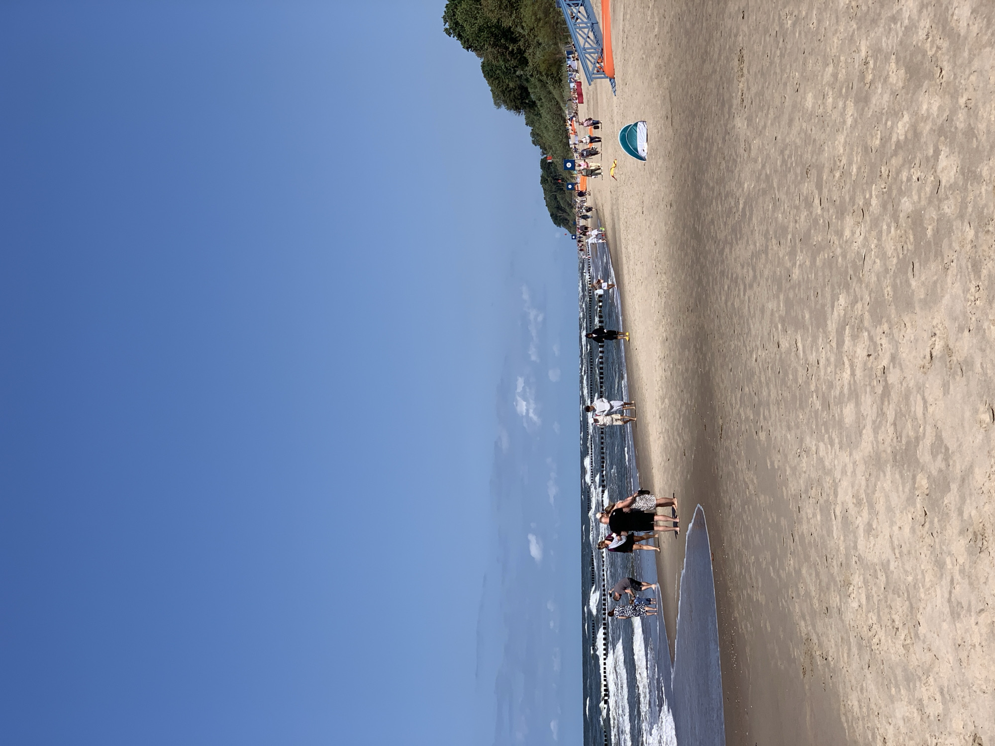 The image depicts a wide, sandy beach in Rewal, Poland, under a clear blue sky. The ocean is calm with moderate waves, and a line of wooden breakwaters stretches across the water. Several people are visible on the beach, some walking along the shore, others near the water's edge.
In the foreground, the sand is textured and appears dry, with some footprints and natural debris. Closer to the water, the sand is wet and darker. A small, light blue and white tent is set up on the beach.
Further down the beach, a group of people are gathered, and in the distance, more individuals are seen strolling. Along the edge of the beach, a densely wooded area with lush green trees forms a natural backdrop. On the right side of the image, a bright orange structure with blue railings, possibly a ramp or seating area, is partially visible. The overall atmosphere is one of a relaxed summer day at the seaside.
