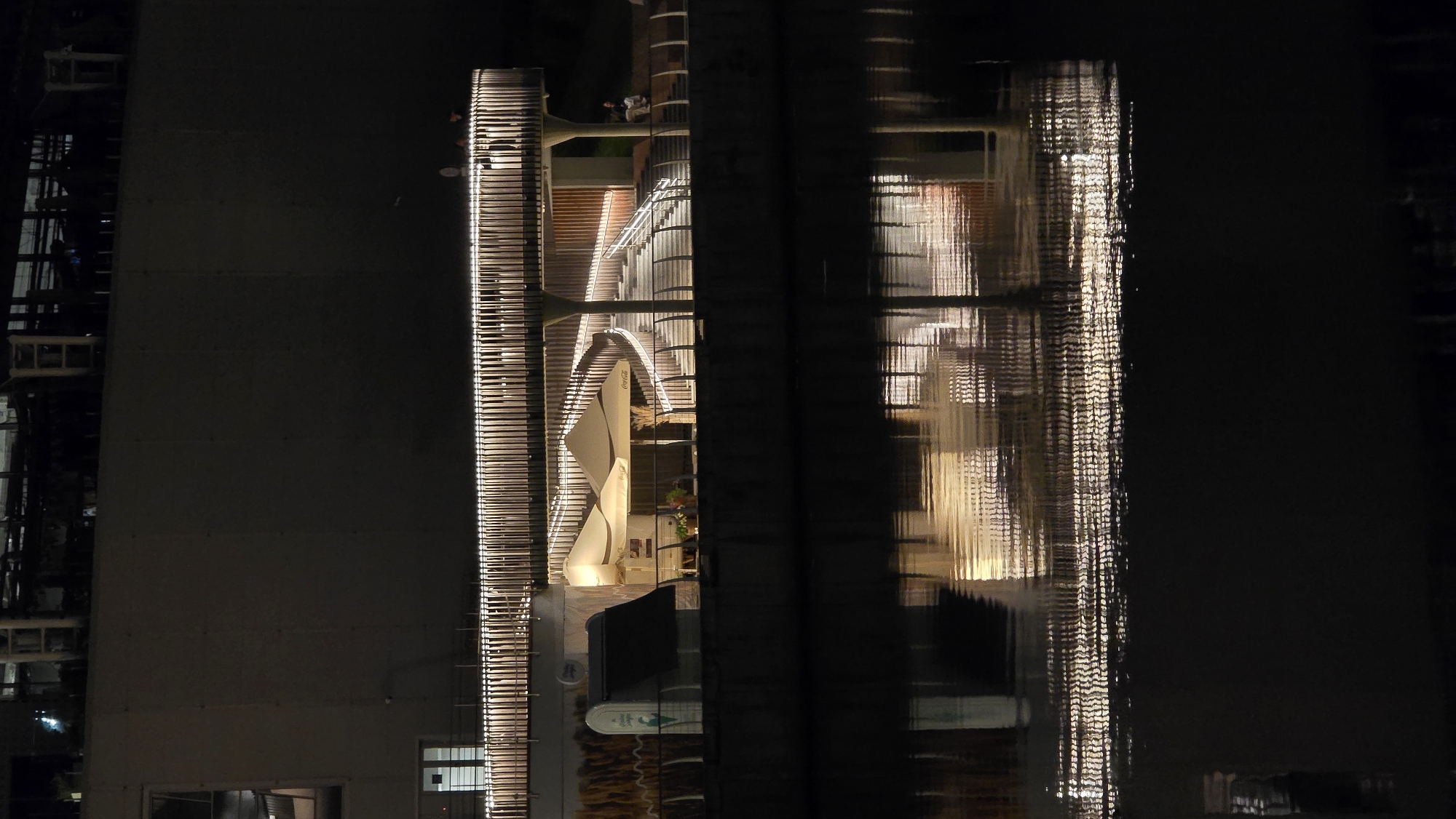 The image depicts a nighttime scene of a modern architectural structure along a waterfront, likely in the city of Kyiv, Ukraine. The structure features a curved walkway or bridge illuminated by a series of vertical, warmly lit posts, creating a striking visual effect against the dark background. Below the walkway, there are at least two large, cream-colored umbrellas, suggesting an outdoor seating area or cafe. A staircase with railings is visible, winding its way up to the upper level. The building behind the walkway is dark and indistinct, with some windows visible at the top. The scene is reflected in the dark water in the foreground, with the lights from the structure creating a shimmering, abstract pattern on the water's surface. Several individuals are subtly visible on the walkway and on a bench in the background, appearing as silhouettes in the dim lighting. The overall impression is one of urban modernity and nighttime ambiance.