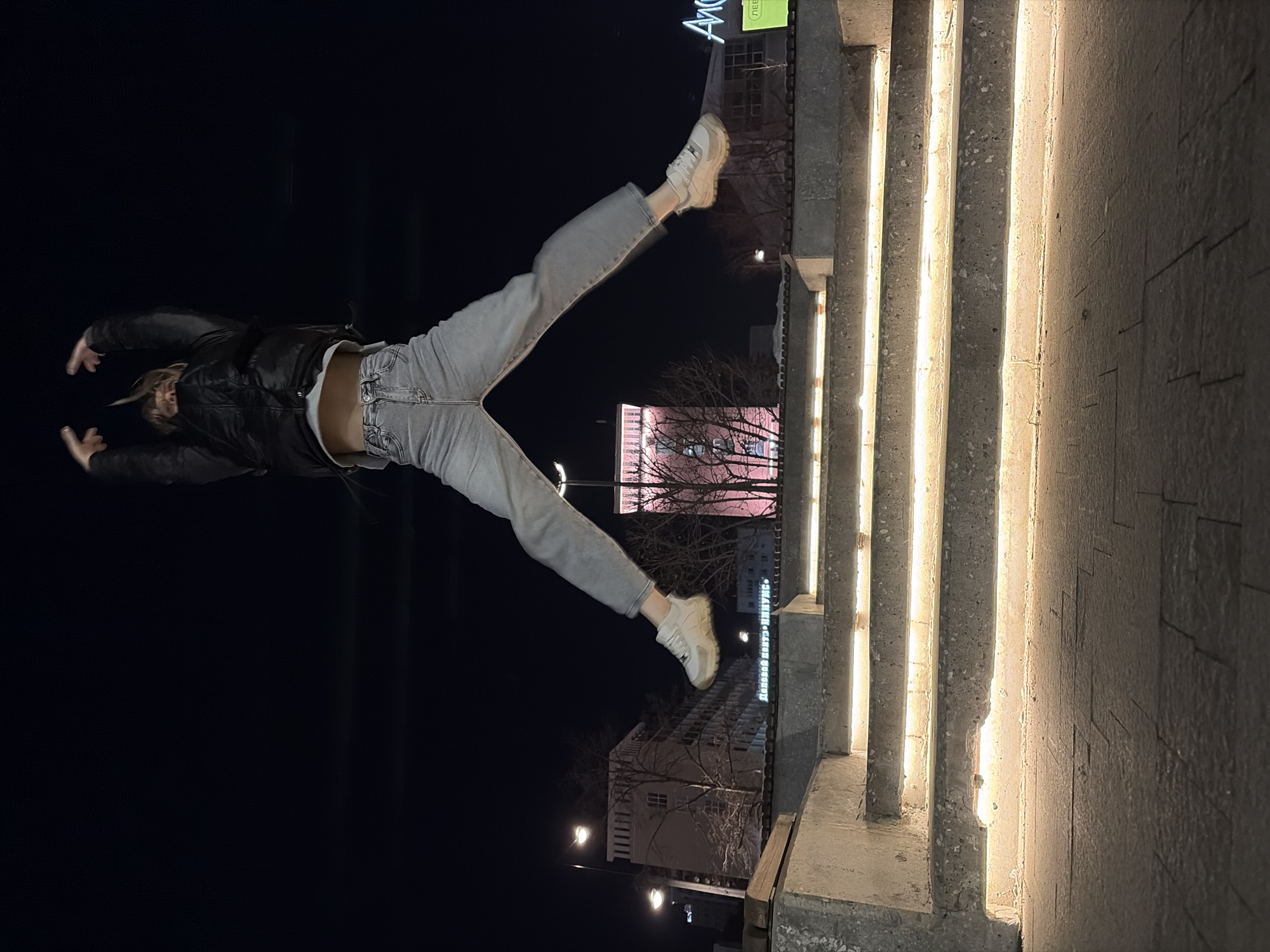The scene depicts a single individual captured mid-jump during nighttime in Perm, Russia. The subject is positioned centrally, elevated above a set of concrete stairs. This individual wears a dark, possibly black, puffer jacket, a light-colored crop top exposing the midriff, light-wash denim jeans with wide legs, and light-toned sneakers. Both arms are raised above the head, and the legs are spread wide. The face is obscured.
In the foreground, a multi-tiered concrete staircase is illuminated by warm-toned linear lights embedded along the underside of each riser. The ground in the immediate foreground consists of dark, rectangular paving stones.
The background reveals an urban environment. To the left, a multi-story building, identified by a visible sign as "Деловой центр «ИРИС»" (Business Center "IRIS"), is present. Two streetlights with multiple fixtures illuminate this area. Centered behind the jumping individual, a distinctive tower, the Perm State Medical University clock tower, is prominently illuminated with pink light. Bare trees are visible in the mid-ground, silhouetted against the dark night sky. To the far right, a partial illuminated sign reads "Апс."