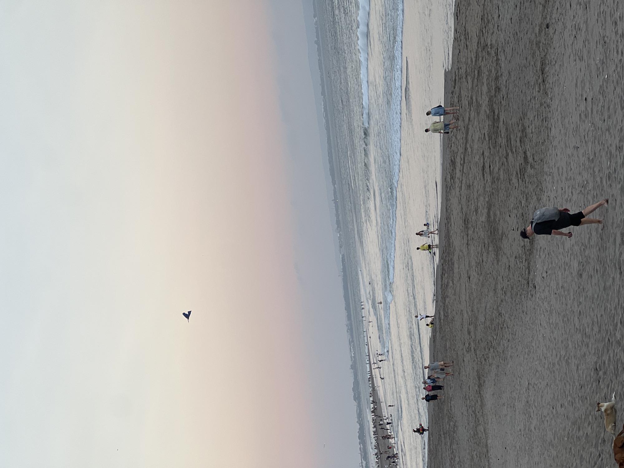 This is a vertical, high-angle photograph of a crowded beach in North Kuta, Indonesia. The image captures a wide expanse of sand and ocean under a hazy sky, likely during sunset due to the warm, soft lighting. Several people are visible on the beach, some walking along the shore, others gathered in small groups. A few individuals are in the shallow water, and waves are gently rolling onto the sand. A single dark kite is flying in the sky on the left side of the frame. The overall atmosphere is relaxed and casual, typical of a beach day. The scene appears to be a popular tourist or local gathering spot, with a long stretch of coastline visible in the background, dotted with distant structures. There is no visible text in the image.