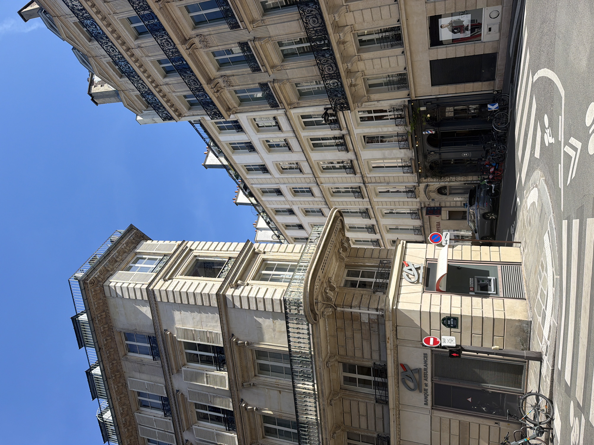 The image shows a street scene in Paris, France. The camera is pointed upwards at an angle, capturing the facades of two buildings. The building on the right side of the frame is a beige, multi-story structure with ornate architectural details, including decorative balconies, window frames, and cornices. A bank branch, labeled "BANQUE ET ASSURANCES," is visible on the ground floor. Traffic signs, including a red stop sign and a bicycle sign, are mounted on a pole nearby. A bicycle is parked in front of the bank. The building on the left side of the frame is a taller, white building with multiple windows and ornate iron railings on the balconies. A shop with a barber pole is also visible along the street, in front of the tall building. A zebra crossing is visible in the foreground, painted on the road. Bicycles are parked near the shop. The sky is blue.