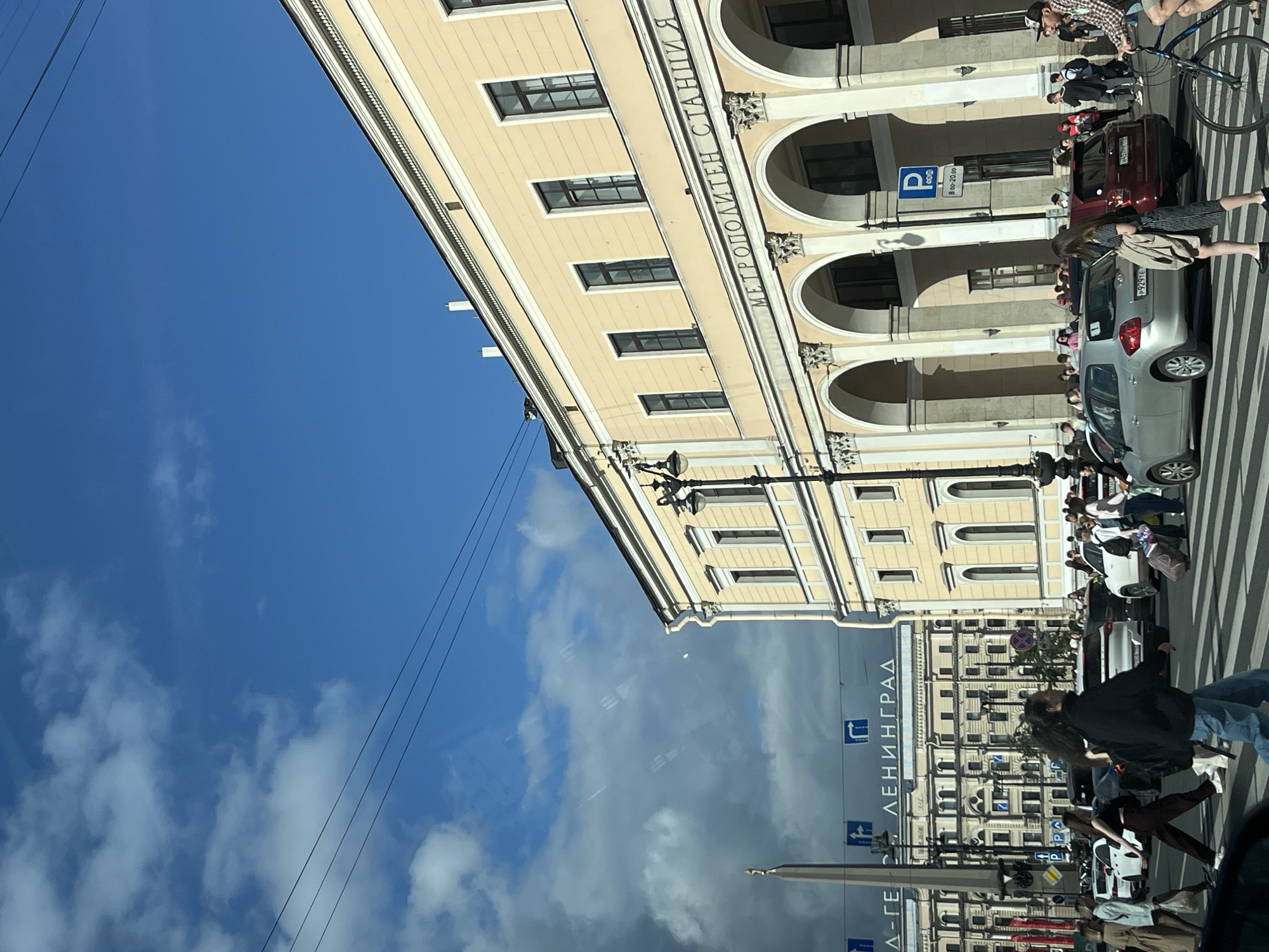 This is a photograph taken from within a vehicle, looking out at a busy city street scene in Saint Petersburg, Russia. The main focus is a large, pale yellow building with arched colonnades and multiple windows, characteristic of classical architecture. Above the arches, there is text in Cyrillic which reads "МЕТРОПОЛИТЕН СТАНЦИЯ", indicating it is a metro station.
In the foreground, a pedestrian crossing is visible with several people crossing. Some are walking, while others are pulling luggage. There are cars stopped at the crossing, and traffic appears to be moving on the street. The weather is partly cloudy with a bright blue sky visible in the upper portion of the image.
In the background, the Obelisk of the Conquerors of the City of Leo is partially visible on the left, a prominent landmark in Saint Petersburg. Further behind, another large building with many windows can be seen, along with blue directional street signs. The overall impression is a bustling urban environment on a clear day.