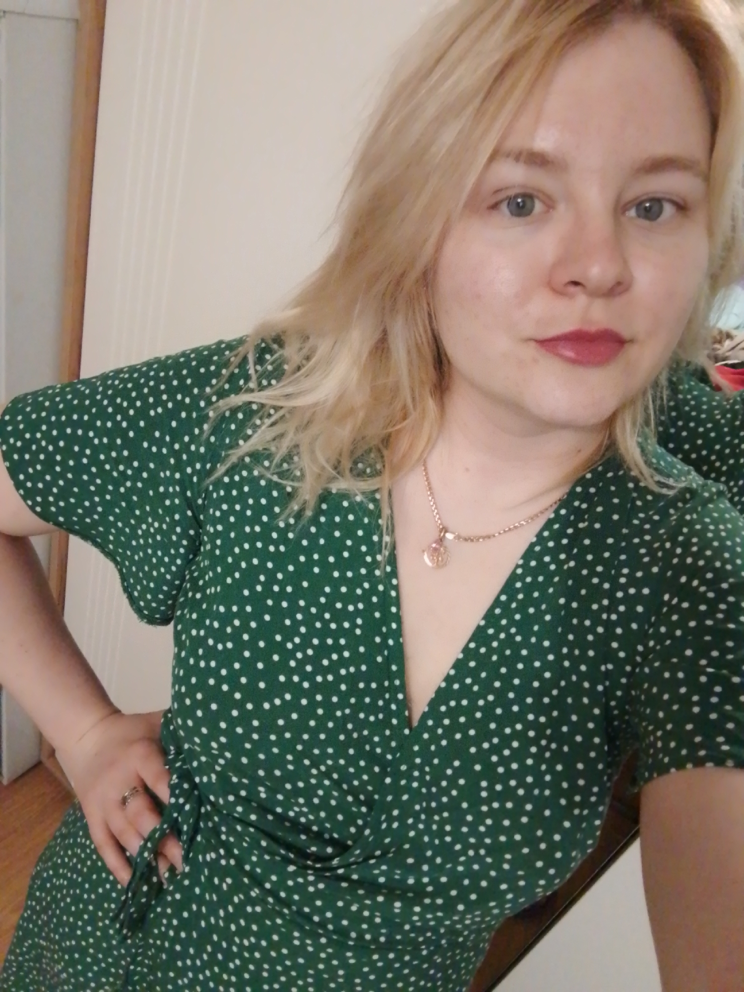 A young woman with blonde hair and blue eyes is posing for a selfie indoors. She is wearing a green wrap dress with white polka dots and a gold necklace. She has a ring on her finger. The background is a white wall with a wooden door frame. The location is not identifiable as Izhevsk, Russia.