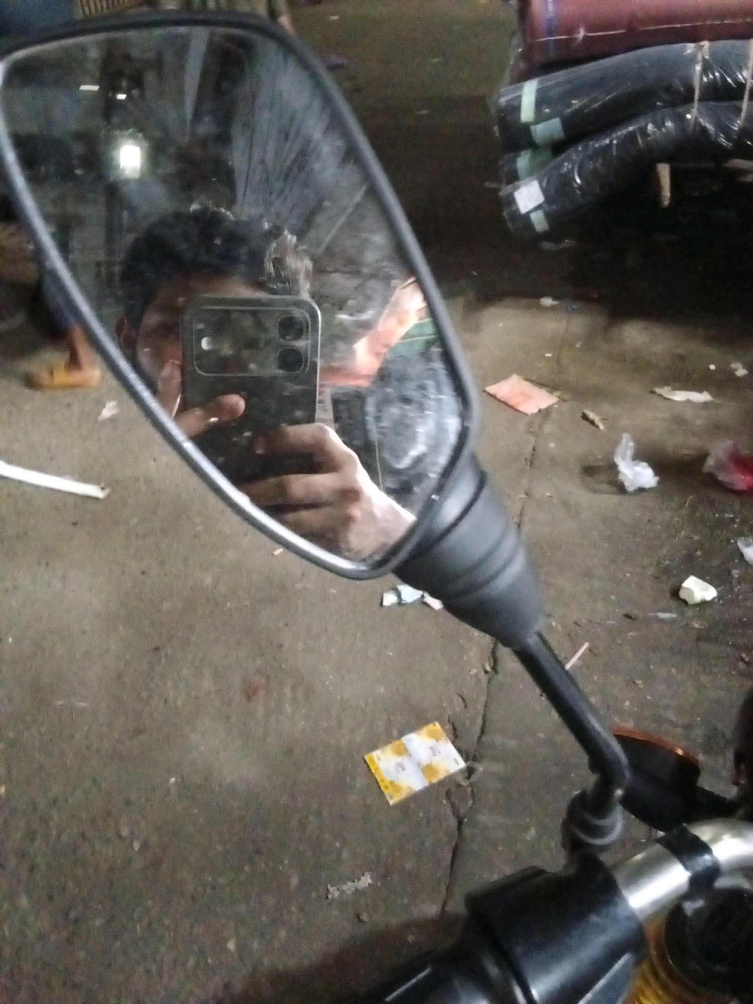 A close-up, low-angle, vertical shot captures a reflection in a motorcycle's rearview mirror. The mirror's surface shows a person's face and upper torso, their hand holding a smartphone used to take the picture. The smartphone has a distinct, triple-lens camera module. Behind the person, the environment appears to be an indoor market or warehouse; stacked rolls of fabric, likely textiles, are visible in the background, wrapped in plastic. The floor is concrete and shows signs of wear and debris, including scattered plastic bags and paper scraps. The overall lighting suggests an interior space, possibly dimly lit or at night. This scene is likely situated in a busy commercial area of Dhaka, Bangladesh, given the commonality of such markets and the presence of motorcycles.