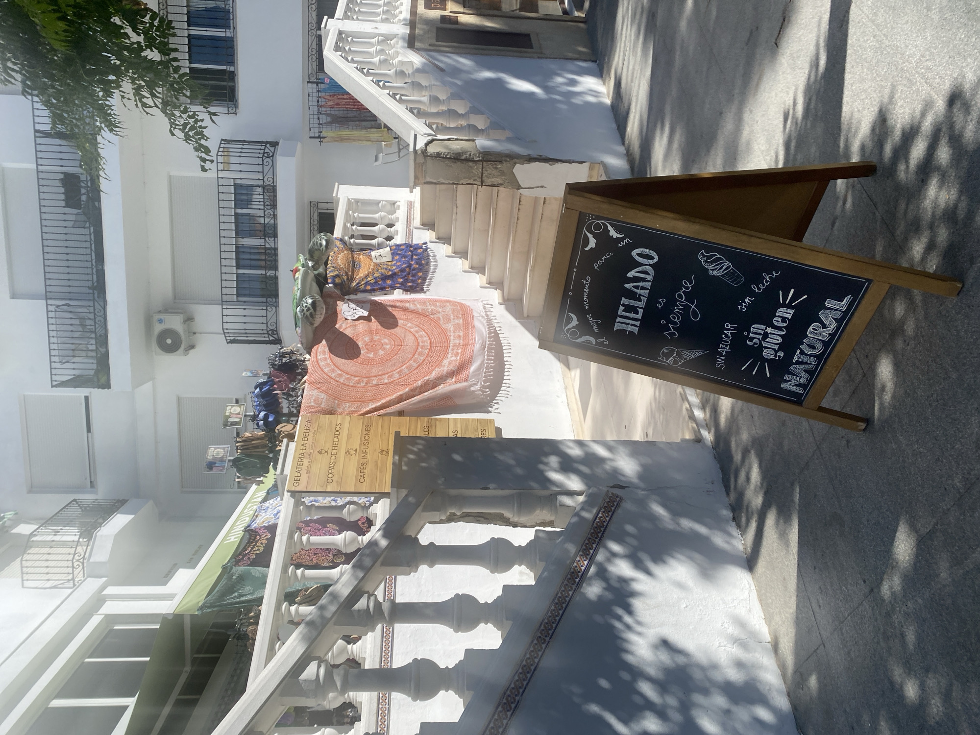 The image captures a bright, sunlit street scene in Conil de la Frontera, Spain. In the foreground, a wooden A-frame chalkboard sign displays handwritten text in white chalk advertising "HELADO" (ice cream) and its qualities: "sin azúcar" (sugar-free), "sin gluten" (gluten-free), and "NATURAL." Drawings of ice cream cones adorn the sign. Behind the sign, a white stone staircase with ornate balustrades leads up to a building. Various colorful fabrics and tapestries are displayed along the staircase and a green awning. The building itself is white with balconies and windows. The ground is paved with grey flagstones, and dappled shadows from trees fall across the scene, indicating it is daytime. There are no people visible in the frame.