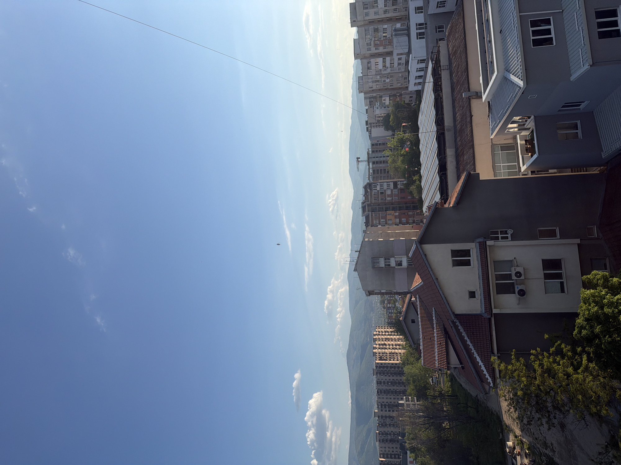 This is an elevated, slightly canted view of an urban area, possibly in Plovdiv, Bulgaria, as suggested. The sky, taking up a large portion of the frame, is bright blue with scattered white clouds. A thin wire or cable stretches across the upper portion of the sky. In the middle ground, there's a line of buildings with varying heights, ranging from single-story structures to tall apartment blocks. Mountains are faintly visible in the distance, appearing hazy due to atmospheric perspective. Close to the right edge of the image, there are houses with red tiled roofs, a gray painted building with an air conditioning unit, and green foliage, which suggests a residential area with trees and vegetation. The lighting is bright, indicating a sunny day. No people or actions are immediately discernible.